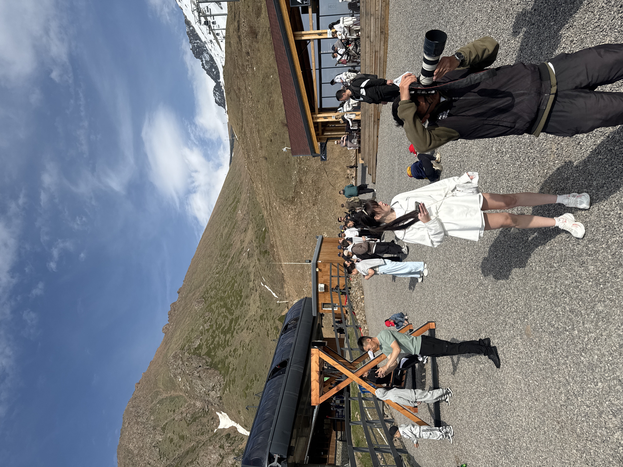 The image depicts a scene at the Shymbulak ski resort in Almaty, Kazakhstan. A man with a professional camera takes a picture of a woman in a white dress. Other people are visible in the background, some on the deck of a building. In the foreground, a man in a green shirt sits on a wooden structure, while others are near a ski lift car. Mountains with partial snow cover form the backdrop under a sky with scattered clouds.