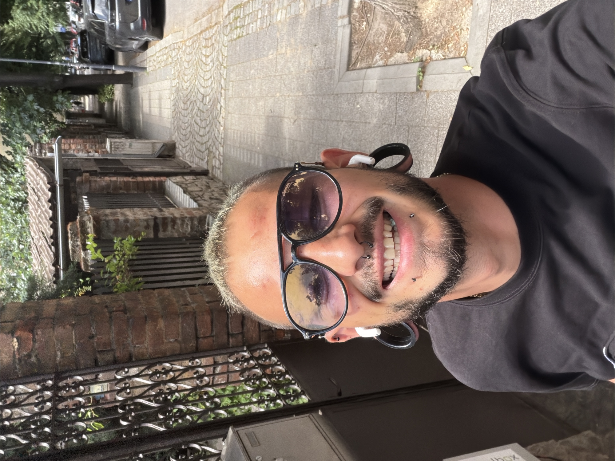 A man with fair skin, a beard, and sunglasses stands on a cobblestone street. He has facial piercings and white earbuds in his ears. A dark-colored t-shirt is visible on his upper body. Behind him is a brick building with wrought iron details. Cars are parked further down the street. The photograph shows the typical cityscape of Plovdiv, Bulgaria, particularly in older neighborhoods with cobblestone streets and brick architecture.