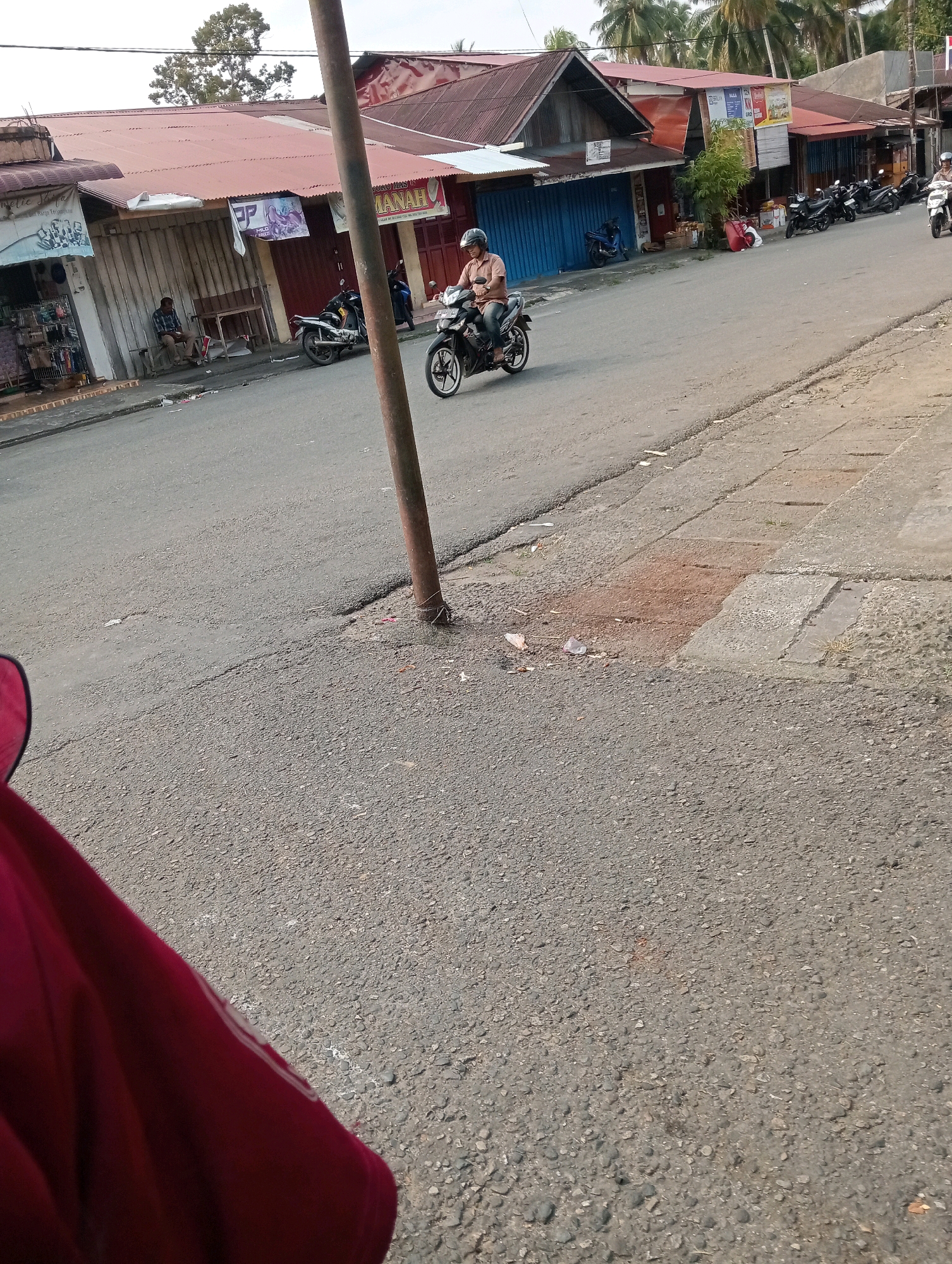 The image depicts a street scene in Pariaman, Indonesia, with a focus on a row of low-rise buildings and the paved road in front of them. A man wearing a helmet is riding a motorcycle on the right side of the road, heading away from the viewer. Several other motorcycles are parked along the sidewalk in front of the buildings. Some of the buildings appear to be shops, indicated by signage and open shutters. A man is seated on a bench in front of one of the buildings. The environment is somewhat cluttered, with debris visible on the roadside. A wooden pole stands prominently in the foreground, casting a shadow. The sky is overcast.