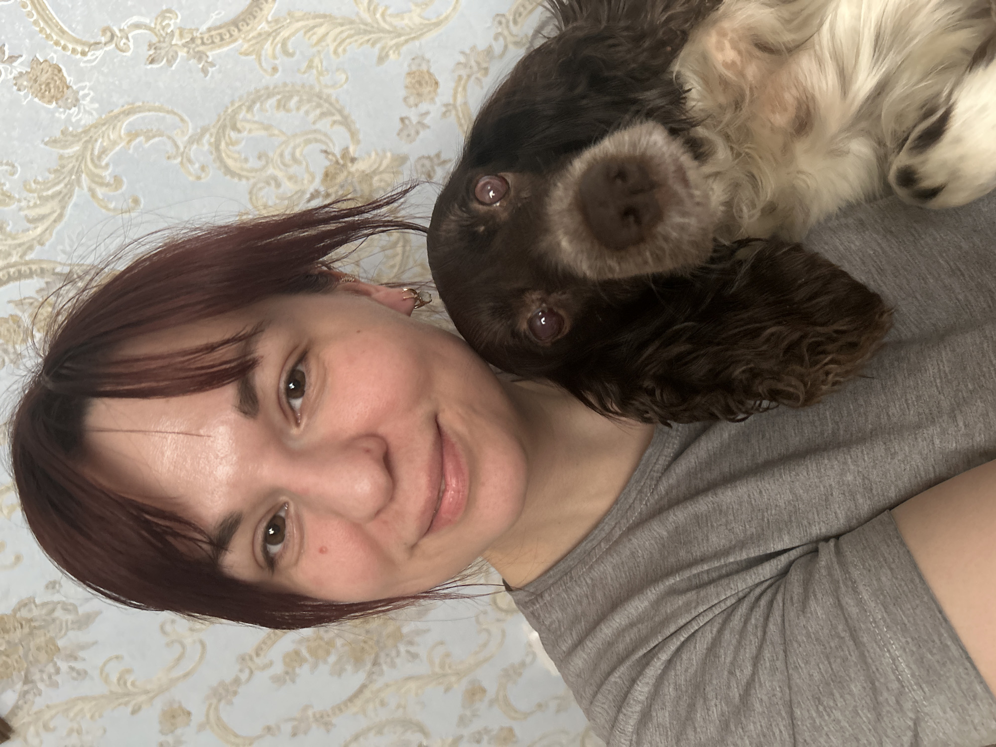The image captures a close-up of a woman and a dog sharing a moment of rest, likely indoors. The perspective is tilted, showing both subjects on their sides.
In the foreground, a woman with medium-length reddish-brown hair and dark eyes is smiling gently towards the viewer. She has a light complexion and appears content. She is wearing a simple grey t-shirt, and a small golden earring is visible near her left ear.
Nestled close to her, partially resting on her, is a brown and white dog, possibly a spaniel breed. The dog's face is prominent, featuring large, dark, expressive eyes and a brown nose, with dark brown fur and white markings on its muzzle and chest. It also looks directly at the camera, appearing calm and curious.
The background consists of a light blue or grey wallpaper adorned with an intricate, ornate pattern in a golden or beige hue, featuring floral and scroll-like designs. The lighting indicates an indoor setting, potentially during daytime or in a well-lit room. There is no visible text in the image. The overall atmosphere conveys a warm and affectionate bond between the woman and her pet.