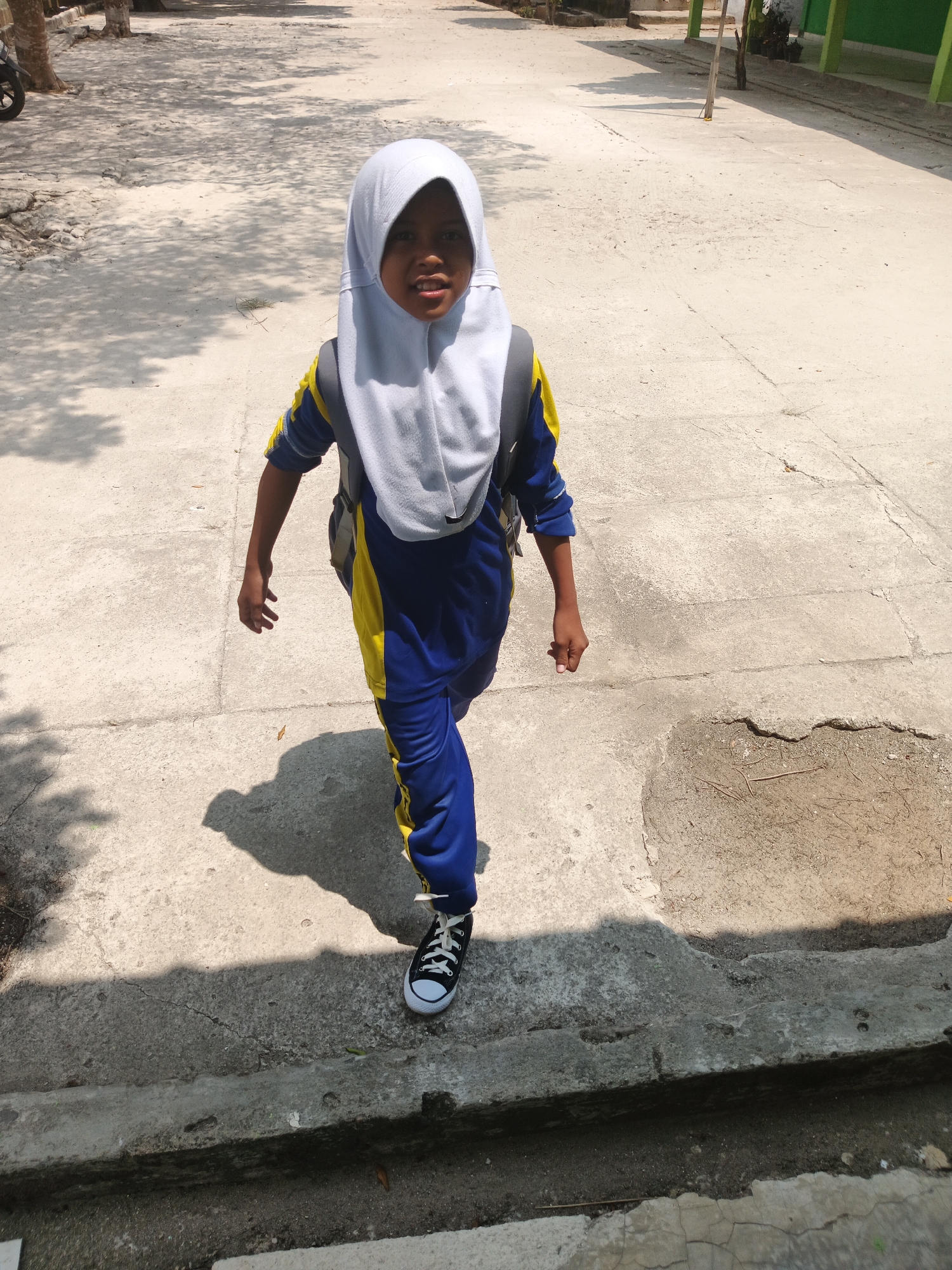 A young child wearing a blue and yellow uniform, a white hijab, and black and white sneakers walks across a concrete path in an outdoor area. The child carries a grey backpack. The ground is a light-colored concrete with some cracks and patches of dirt. In the background, there are shadows of trees and the outline of a green building. The scene appears to be an open area, possibly a schoolyard or public space, in Rejo Basuki, Indonesia.