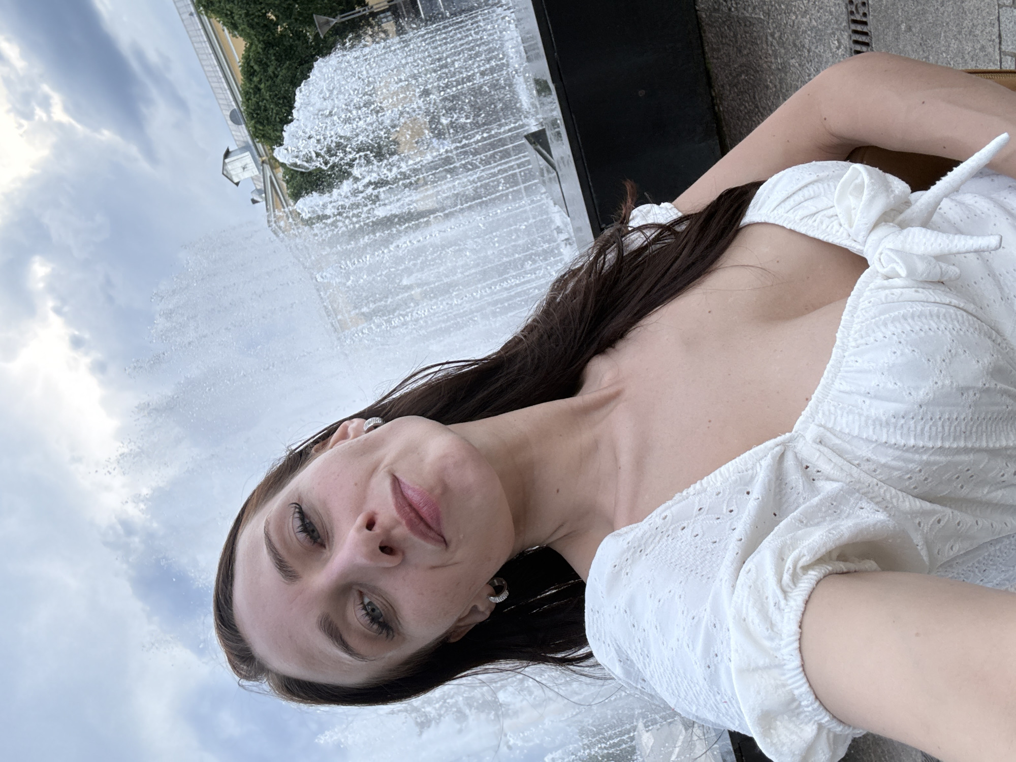 A woman with long, dark hair and fair skin, wearing a white eyelet top with a sweetheart neckline and a bow detail, poses for a selfie in front of a large fountain in Saint Petersburg, Russia. The fountain's water is actively spraying. In the background, a yellow building with a grey roof is visible behind trees. The sky is cloudy.