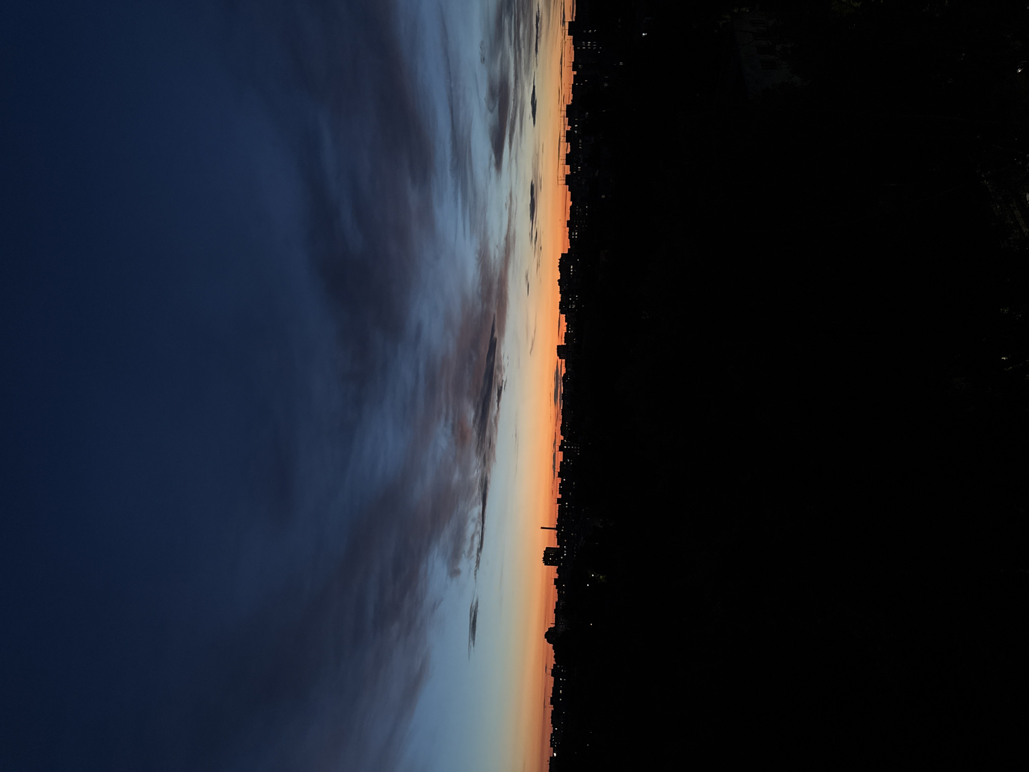 The image captures a dusky skyline of Nizhny Novgorod, Russia, during twilight. The upper portion of the frame is dominated by a deep blue sky, transitioning to a gradient of lighter blues and then a vibrant orange band near the horizon. Wispy, elongated clouds stretch across the sky, catching the last rays of the setting sun and appearing in shades of gray and pink.
Silhouetted against the colorful sky is the cityscape, rendered in deep black due to the low light conditions. Numerous buildings of varying heights are visible, their forms indistinct. Lights are scattered throughout the urban landscape, appearing as small, bright points against the dark buildings, suggesting that some of the city's inhabitants are already indoors. The foreground is a dense, solid black mass, obscuring any details of the ground level. The overall impression is one of a city settling into night, with the dramatic hues of the sky providing a stark contrast to the dark, anonymous structures below. There are no discernible people, specific objects, or actions depicted in this scene.