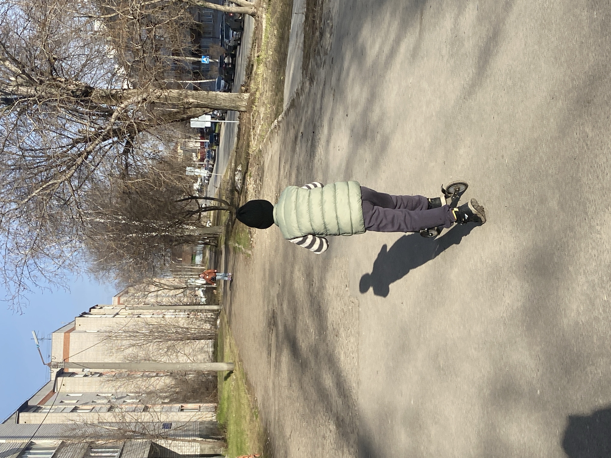 A person is lying on a skateboard on a sidewalk in Kazan, Russia. The person is wearing a black beanie, a light green puffer vest with a striped shirt underneath, and gray pants with black shoes. The sidewalk is gray concrete and runs alongside a road with trees and buildings. The trees are mostly bare, suggesting it is either late autumn or early spring. Buildings and cars are visible in the background. The weather appears sunny.