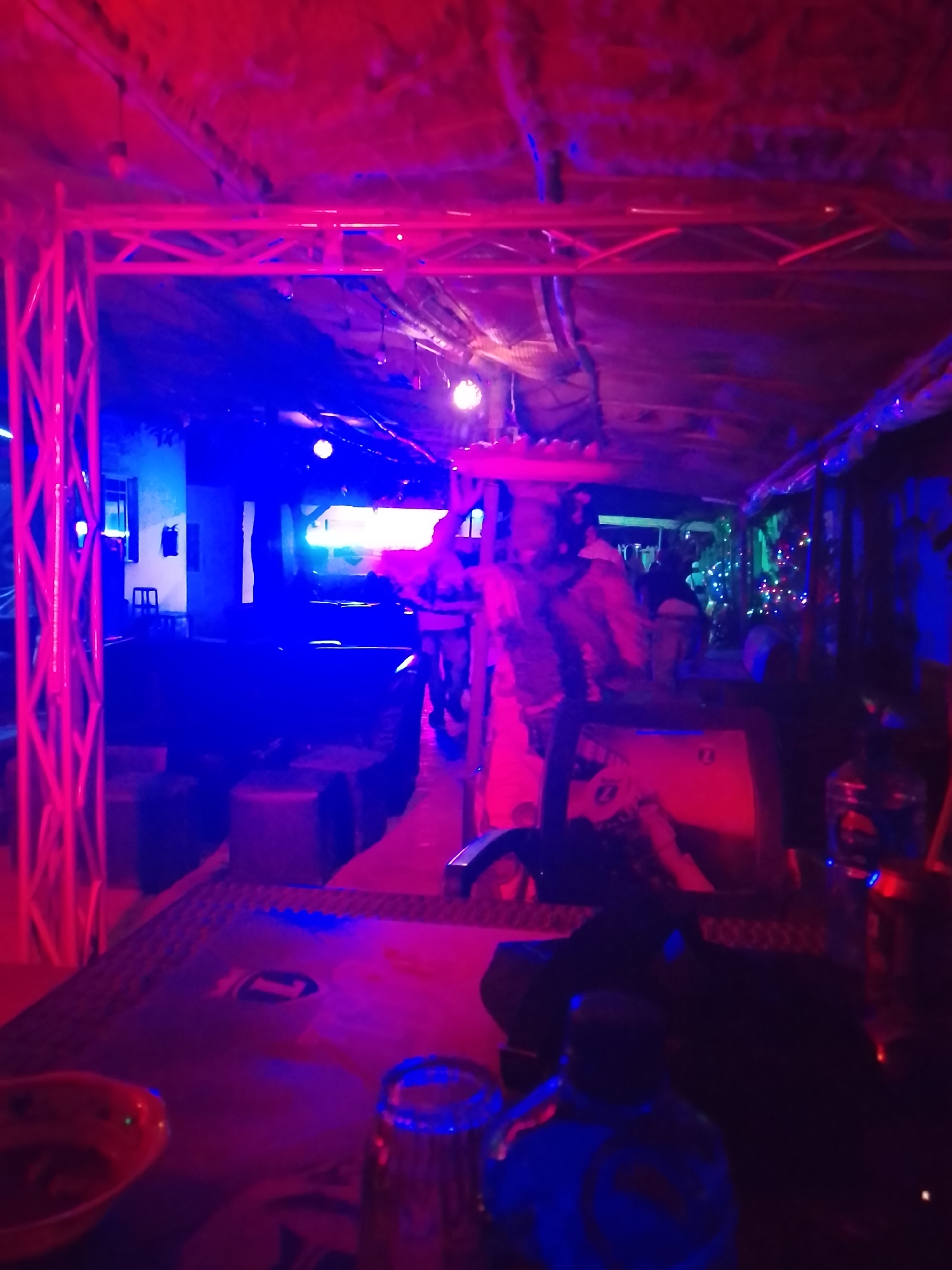 Captured indoors in Unknown, Unknown, the scene is bathed in vivid red and blue lighting. The focus is on the interior of what seems to be a bar or club. A structural frame, illuminated in red, spans across the upper part of the image. The dimly lit furniture includes seating arrangements and tables. Toward the center, figures are seen moving or standing, although details are obscured by the lighting. The background shows lights and possibly decorations, suggesting an entertainment venue. In the foreground, objects on a table are visible, including a glass, a bottle, and other items. The scene indicates nightlife activity in an urban setting.