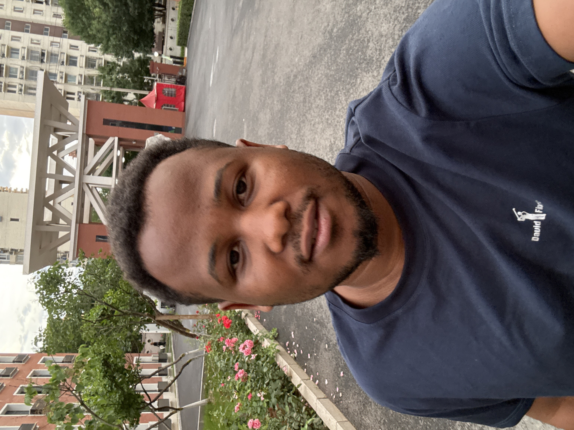 A medium shot captures a Black man in a navy blue t-shirt standing outdoors. The man faces the camera with his head slightly tilted. He has short, dark, curly hair and a neatly trimmed beard. Behind him, a paved pathway runs alongside a flower bed with pink and red roses. Beyond the flowers, trees with green foliage are visible, and further in the background, a modern building with a reddish-brown facade and white trim stands, featuring a large, open-air architectural structure with a white triangular roof. To the right, a tall residential building with many windows can be seen. A small red tent or booth is situated near the entrance of the building. The ground appears to be asphalt or a similar dark paving material. The sky is overcast. The image is a selfie, suggesting the man is taking the photograph himself. The context of Daxing District, China, is provided as the location.