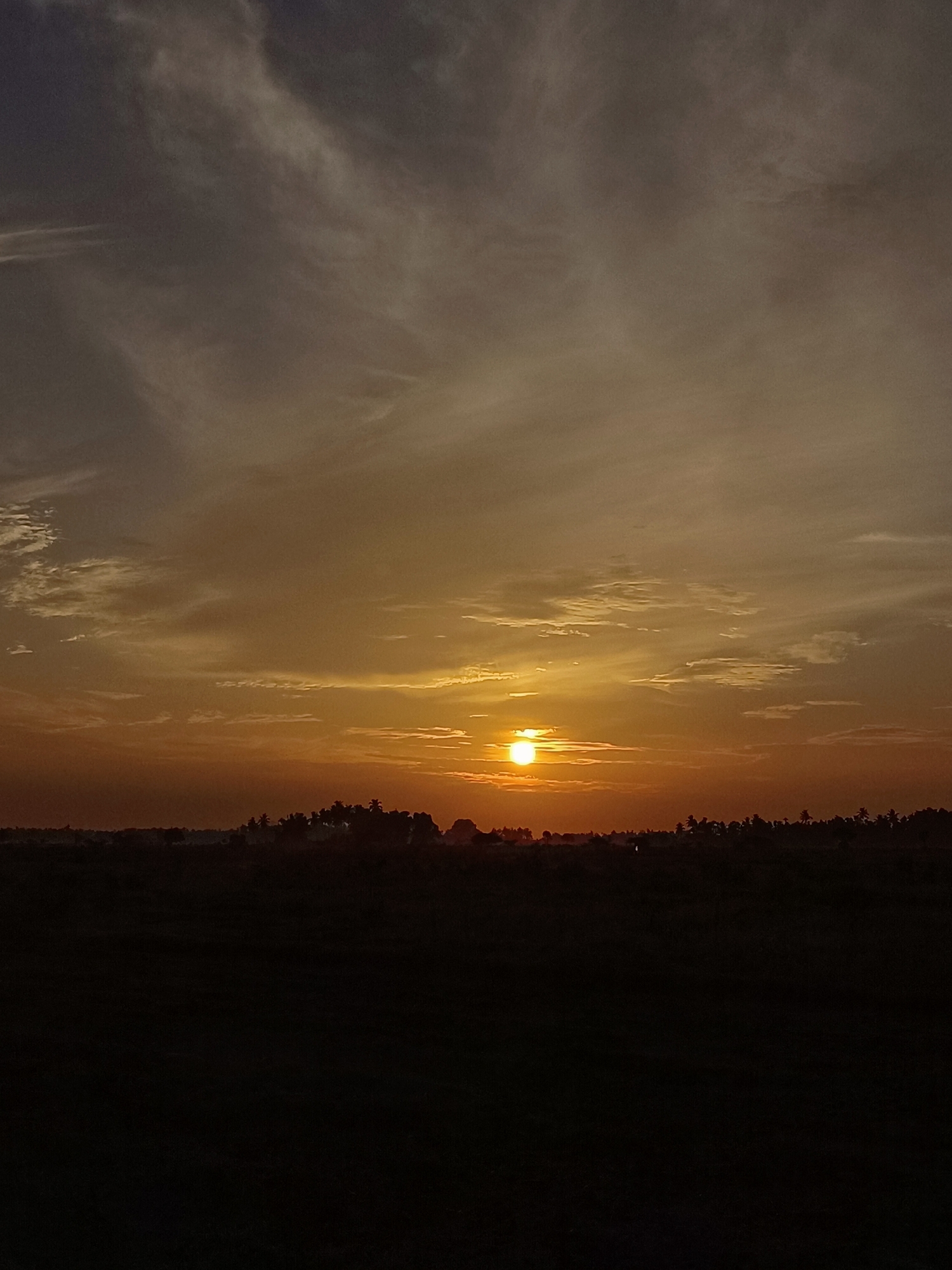 The image depicts a sunset in Meunasah Pupu, Indonesia. The sun is a bright orb low on the horizon, casting a warm, orange glow across the sky, which is filled with streaks of clouds in varying shades of grey and orange. Silhouetted against the vibrant sky is a dark, horizontal band of land, likely representing fields or a low-lying area. On this land, a line of trees, possibly palm trees given the tropical setting, creates a jagged, dark edge against the illuminated sky. The foreground is almost entirely black, suggesting it is a dark field or ground area, with minimal detail visible. The overall impression is one of a serene, rural landscape at dusk.