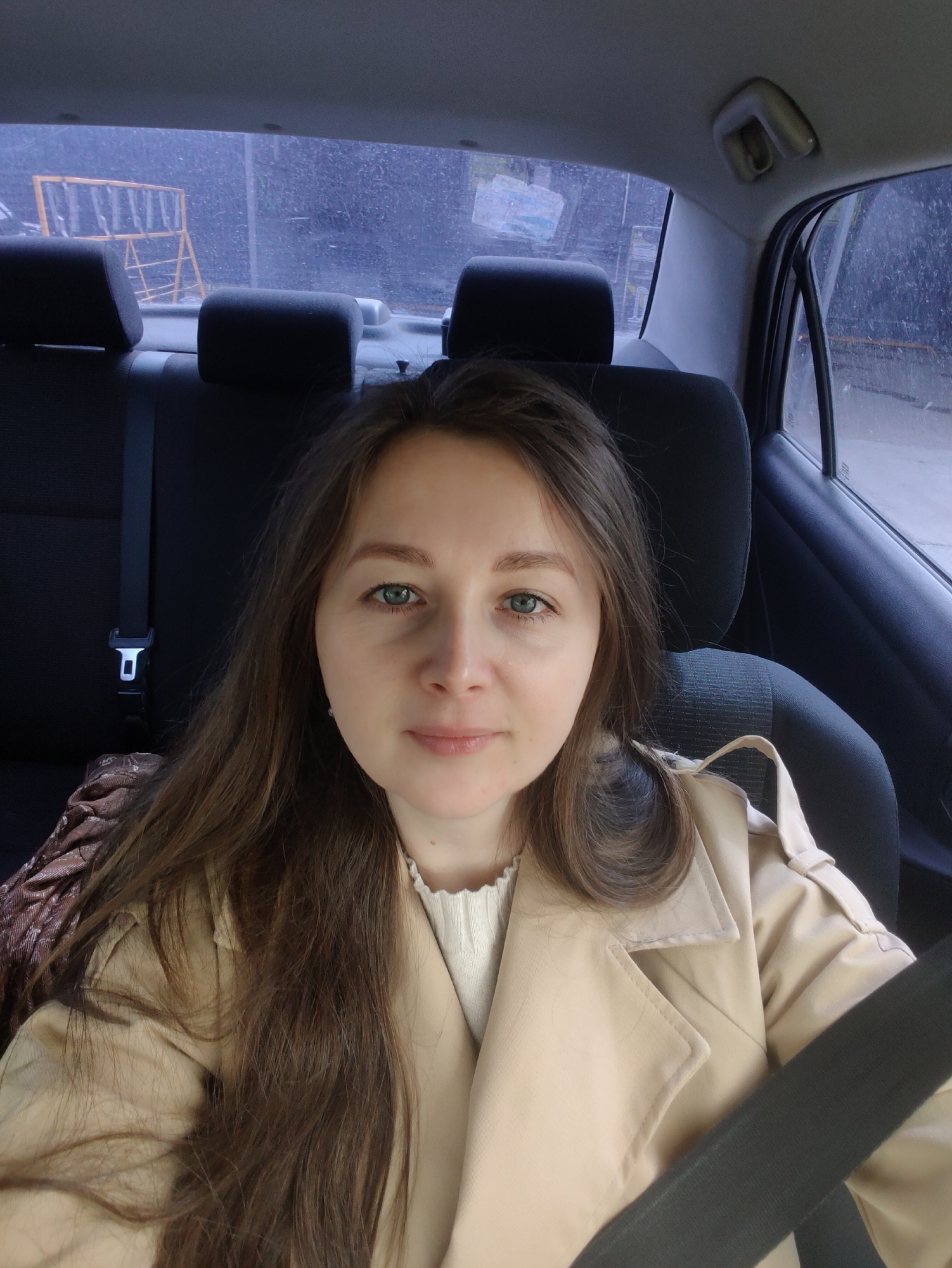 A medium shot depicts a woman with shoulder-length brown hair and light eyes in the back seat of a car. She is wearing a light-colored trench coat and a white top. A black seat belt crosses her chest. The car seats behind her are black. Outside the rear and side windows, it appears to be a gray, overcast day with a blurred view of urban elements. A yellow metal object that resembles a barricade is visible outside the rear window. The city is Unknown, Unknown.