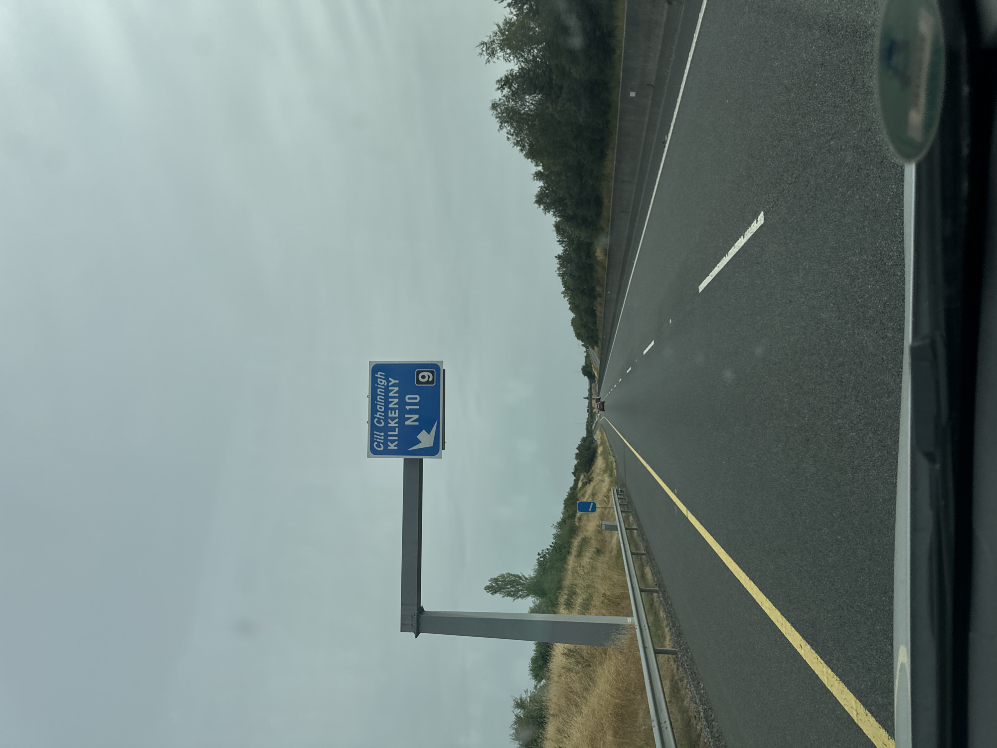 The image depicts a highway scene under an overcast sky. A blue overhead road sign is visible, indicating "Cill Chainnigh KILKENNY N10 9" with an arrow pointing left. The road is asphalt with yellow and white lines marking the lanes. Guardrails and dry grass line the left side of the road, with trees and vegetation in the background on both sides. A few cars are visible in the distance on the highway. The photograph appears to be taken from the perspective of a vehicle driving on the highway. This scene is consistent with a location on the N10 road leading towards Kilkenny, Ireland, as indicated by the signage.