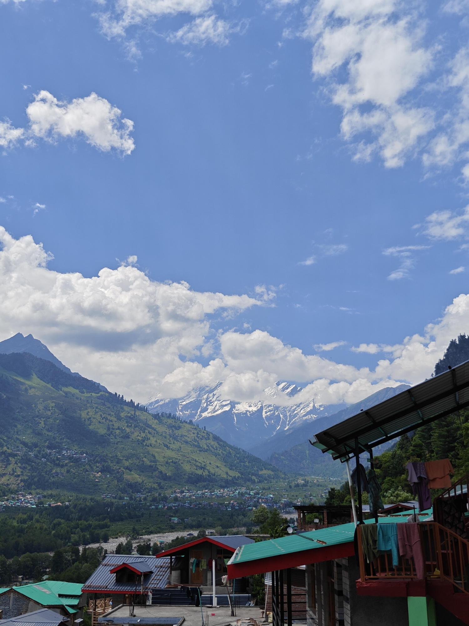 This is a vertical, eye-level, outdoor photograph taken in Vashisht, India, featuring a scenic mountain landscape with a village and several structures in the foreground. The sky is a bright blue with scattered white cumulus clouds. In the background, majestic snow-capped mountains rise, their peaks sharply defined against the sky. The slopes of the mountains are covered in lush green vegetation and dotted with small houses, indicating a village or town nestled within the valley.
In the foreground, several buildings with red and green corrugated metal roofs are visible. One building on the right has a balcony with clothes hanging out to dry on a line. The structures appear to be guest houses or homes, typical of a tourist area. The overall scene conveys a sense of tranquility and natural beauty.