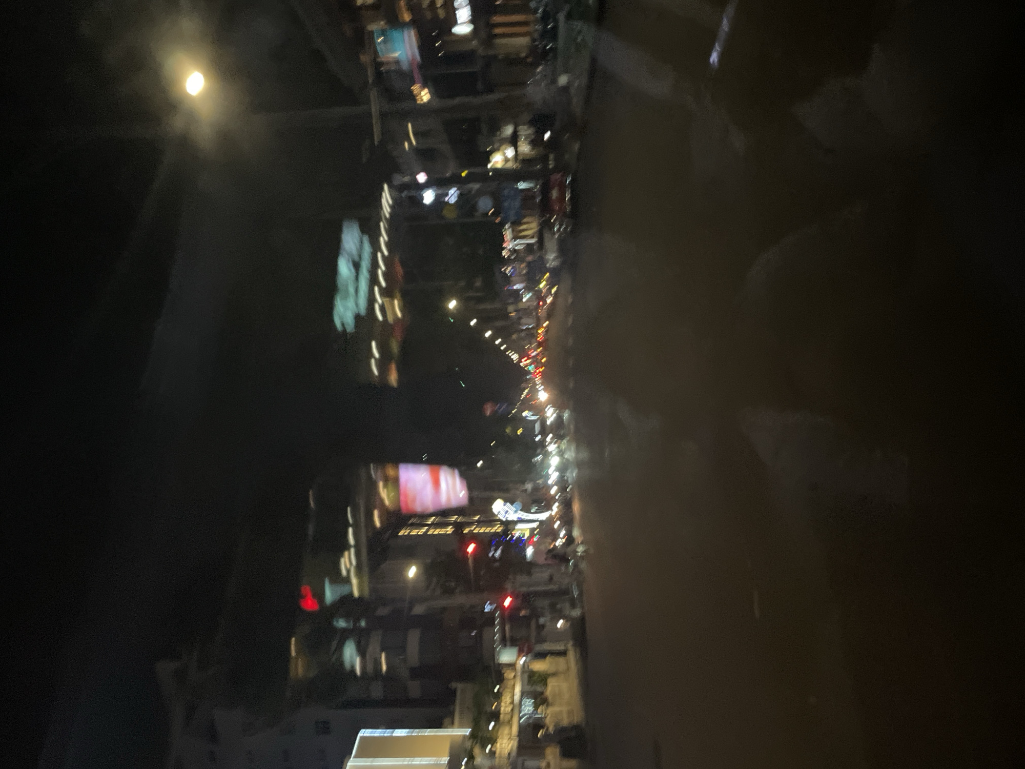 The image depicts a street scene at night in the city of Thủ Đức, Vietnam. The perspective appears to be from a moving vehicle, resulting in a blurred and streaky effect. The road is visible in the foreground, reflecting the ambient lights. On either side of the street, buildings and shops are illuminated with a variety of lights, including signs and building facades. Traffic lights are visible, showing red and other colored signals. Some vehicles are present on the road, contributing to the streaky light patterns. The sky is dark, with a bright, circular light source, possibly a streetlight or the moon, partially obscured by clouds or haze. The overall impression is one of a busy urban environment at night.