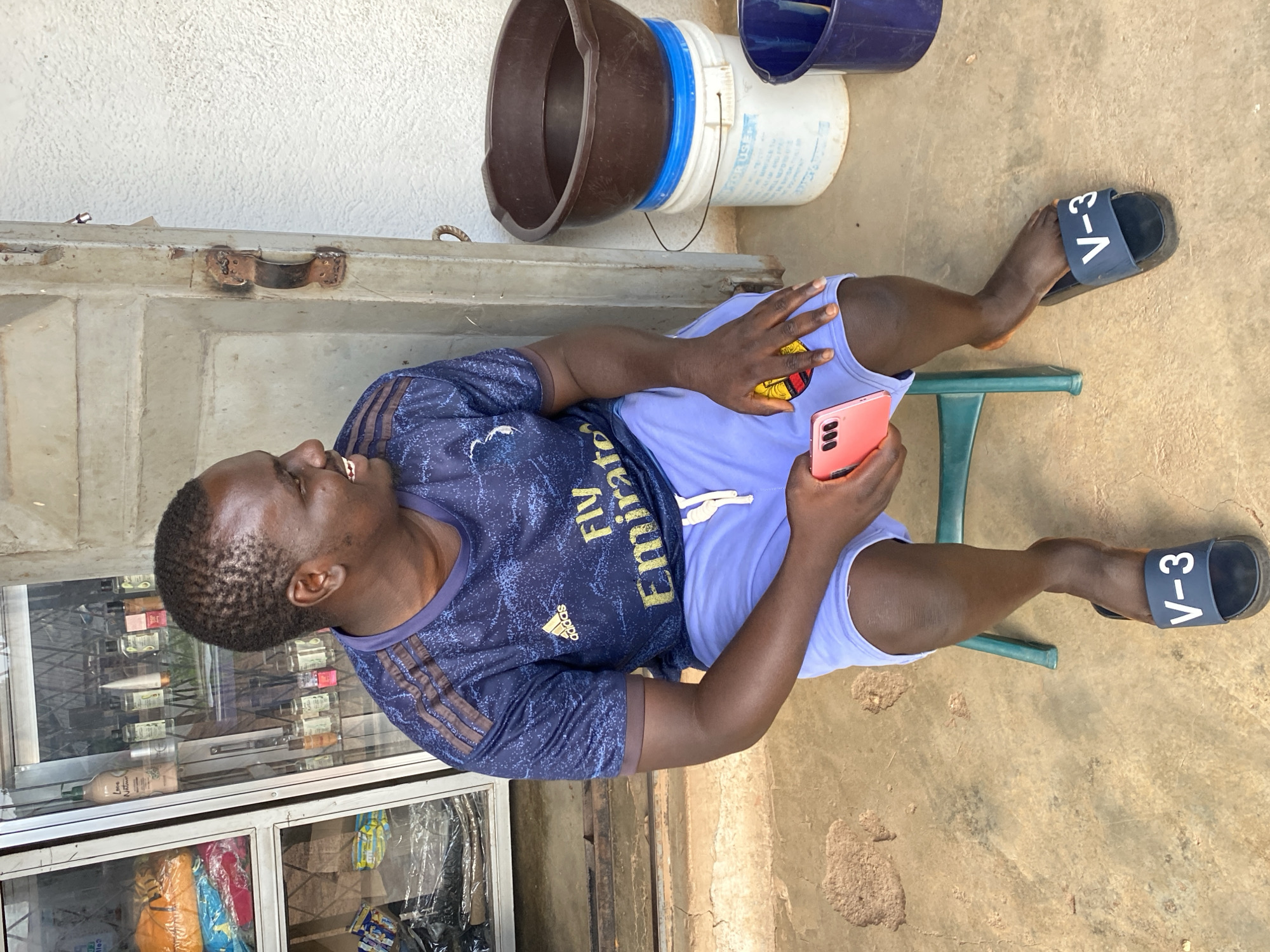 A dark-skinned man sits on a green plastic stool in front of a shop in Bukuru, Nigeria. He is wearing a dark blue Adidas t-shirt with gold stripes on the sleeves and "Fly Emirates" printed across the chest. He also wears light blue shorts and dark blue slides with "V-3" printed on them. He holds a pink smartphone in his right hand. Behind him, a glass display cabinet in the shop contains various products, including bottles and folded clothing. To his right, there are two buckets, one blue and one white, and a large brown basin filled with dark liquid. The ground is a dirt-like surface with some debris. The wall behind the shop is rough and white-washed.