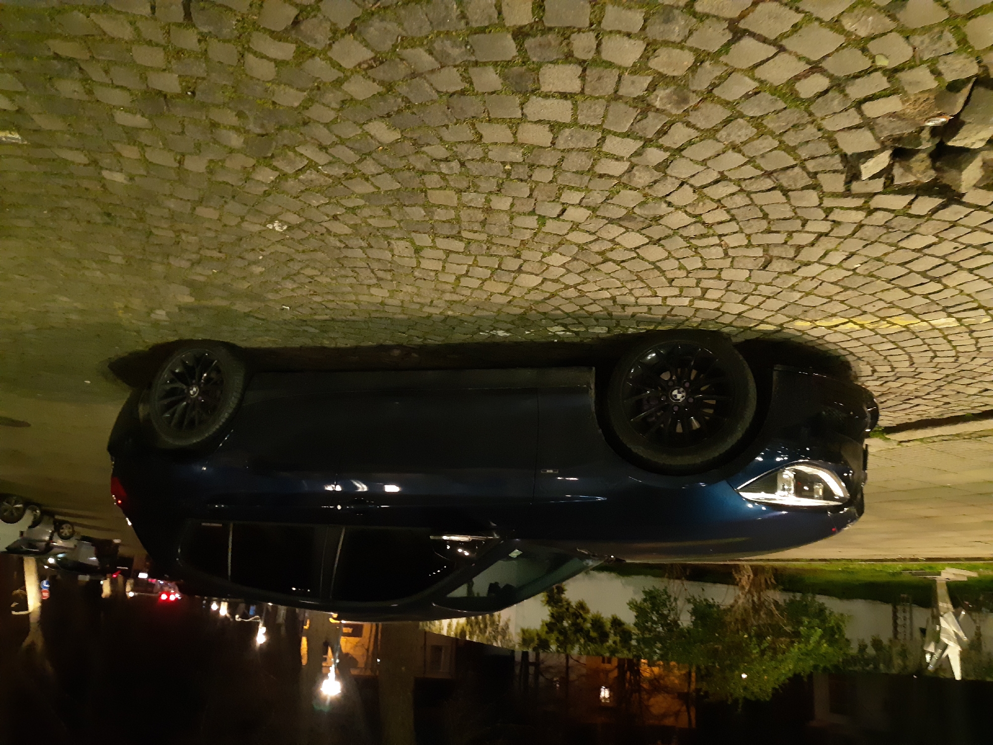 A dark blue BMW is parked on a cobblestone street in Plovdiv, Bulgaria, at night. The cobblestones appear to be arranged in a curved pattern. Other cars are parked in the distance, and the lights from those cars are visible. Trees and buildings line the street, and their reflections can be seen on the wet street surface.