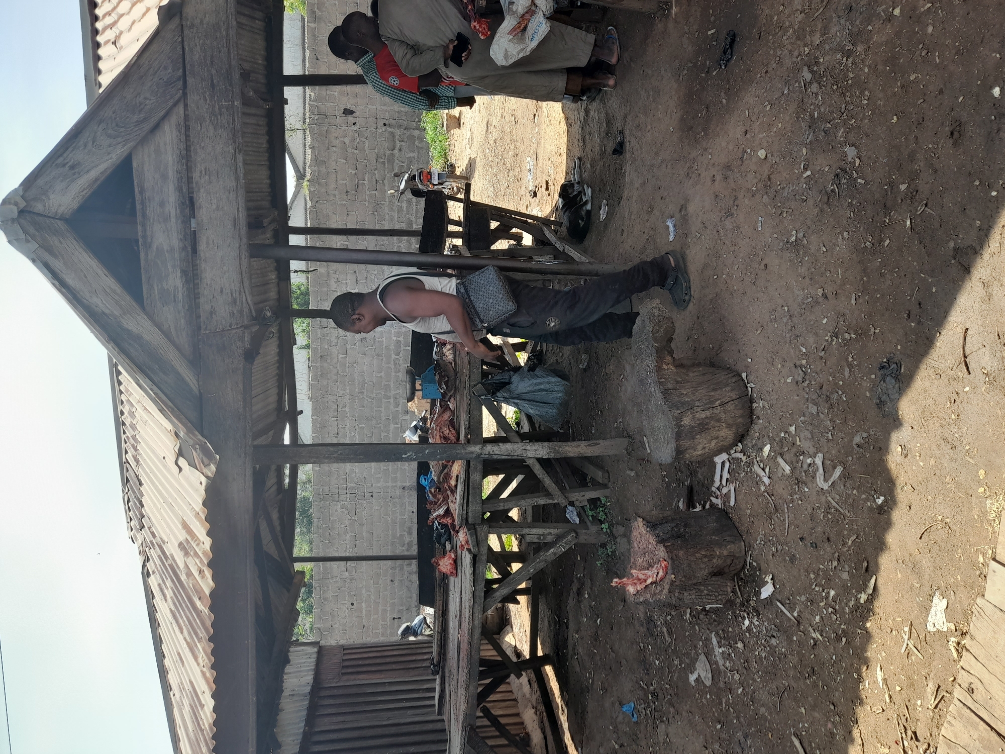 This image depicts an outdoor market scene in Kakuri Gwari, Nigeria, likely a butcher's stall. A man stands near a wooden table laden with raw meat. He wears a white tank top, dark pants, and a black crossbody bag. To his right, two other men are visible, one in a red and green plaid shirt and another in a tan robe. A motorcycle is parked behind the table. The ground is dirt and littered with debris, including bones. Two large wooden stumps are present in the foreground, one with meat on it. The market is covered by a corrugated metal roof supported by wooden posts. A brick wall forms the background. A black and white dog is lying on the ground near the table.