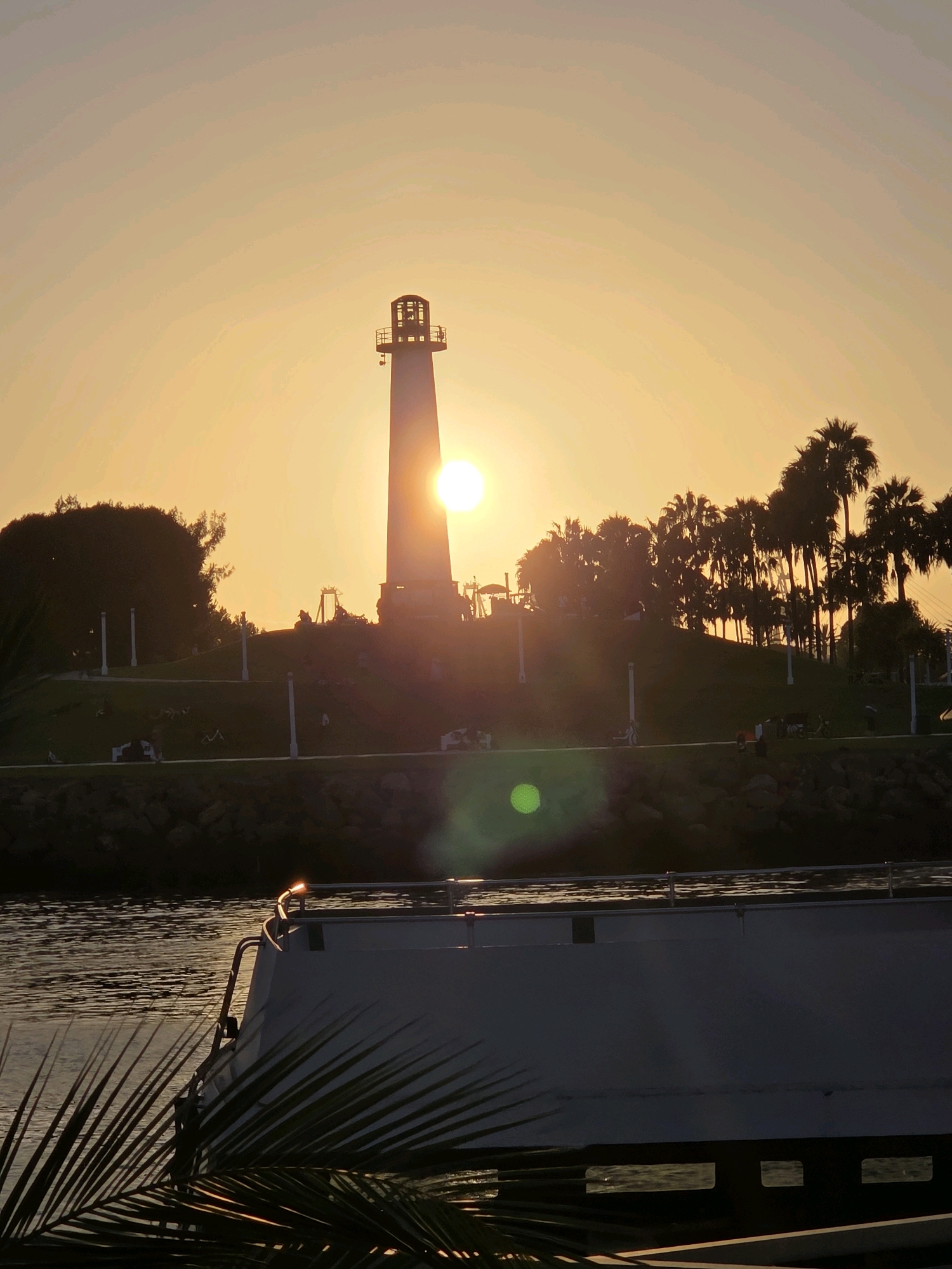 A lighthouse stands silhouetted against a bright orange sunset, partially obscuring the sun. The lighthouse is on a grassy hill populated by palm trees and other foliage. In the foreground, the dark silhouette of a boat is visible on the water, with a palm frond in the lower left corner. The scene appears to be a waterfront area, possibly a harbor or marina. The image is set in Garden Grove, United States of America, though the specific location within the city is not identifiable. There are scattered figures of people on the hill and on the boat, but their activities are not discernible due to the backlighting. The water has ripples and reflects the warm hues of the sunset.