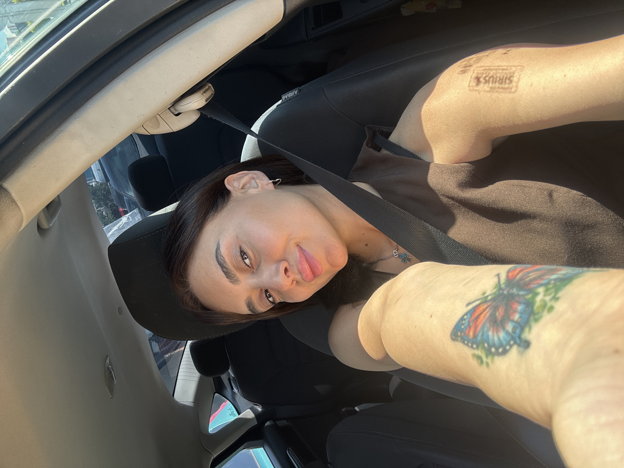 A young woman with dark hair and brown eyes is taking a selfie inside a car. She is wearing a dark brown top and a seatbelt. On her left arm, she has a tattoo of a butterfly. On her right arm, she has a tattoo that appears to be a logo with text. She is smiling and looking at the camera. The interior of the car is visible, including the seats and the ceiling. Sunlight is streaming in through the windows, casting shadows on the woman's face and arms. The background outside the car is blurred, but appears to be a city street with buildings and trees. The image appears to have been taken during the daytime.