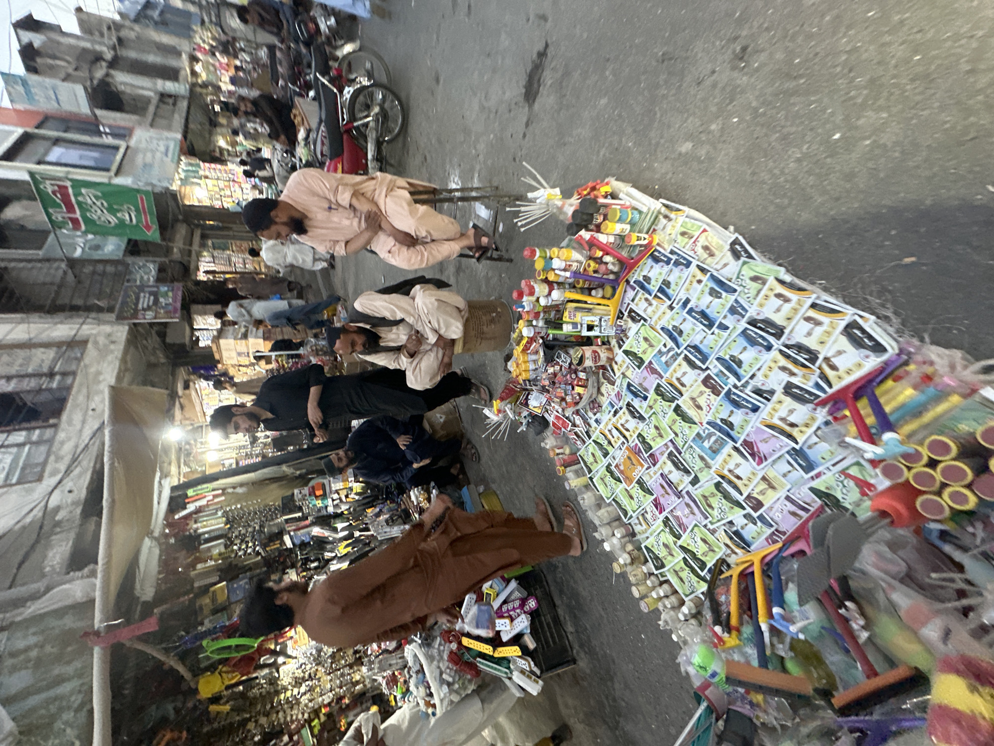 The image depicts a street scene in Quetta City Tehsil, Pakistan. Several men are sitting or standing near various stalls or displays of goods. One stall displays a large collection of small, identical cards, alongside bottles, tubes, and brushes. Other stalls appear to sell a variety of goods, including electronics and household items. The street is bustling with activity, and buildings line the road. A motorcycle is parked nearby. The overall atmosphere suggests a busy marketplace or commercial area.
