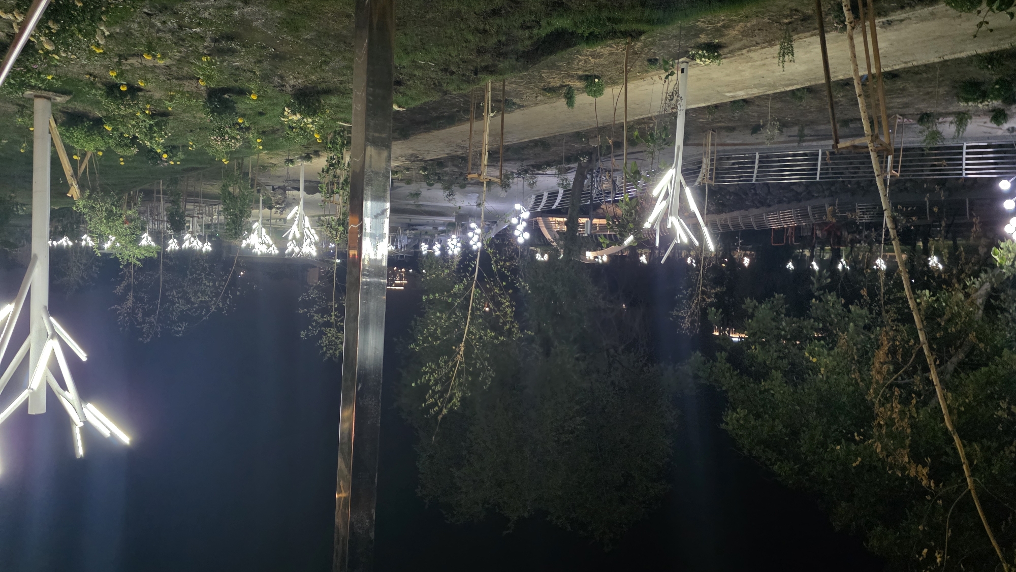 This is a nighttime, low-angle shot of a park in Osh City, Kyrgyzstan. The park is illuminated by modern, tree-shaped light fixtures with white LED branches. A pathway runs through the park, bordered by trees and manicured grass. In the background, a bridge spans across a body of water. The scene suggests a public recreational area, possibly a promenade or a garden, designed for evening use. The lighting creates a surreal and enchanting atmosphere. There are no people visible in the frame. The overall environment is clean and well-maintained, indicating a modern urban park.