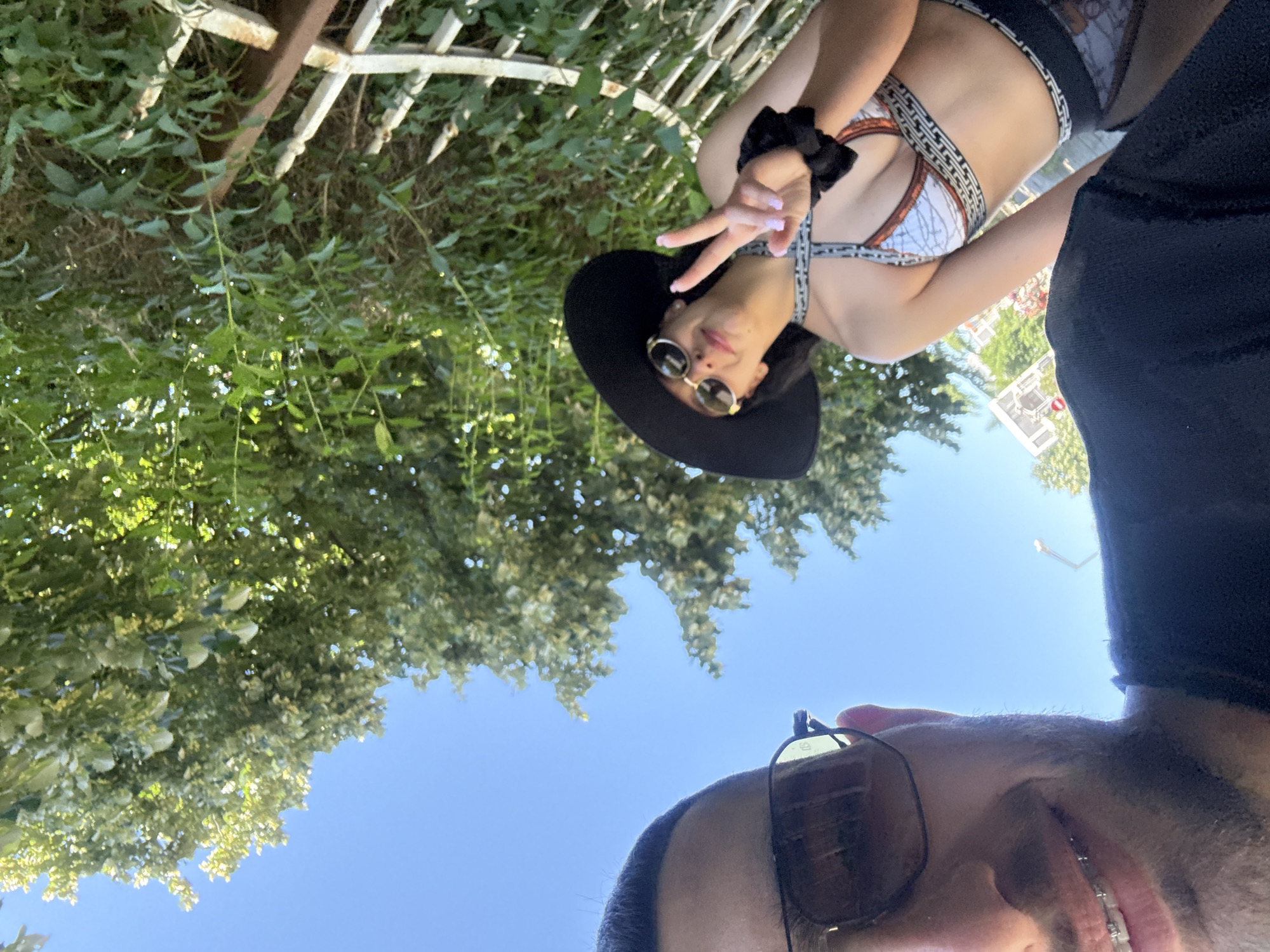 In Nesebar, Bulgaria, a low-angle selfie captures a man in the foreground, his face partially obscured by large sunglasses with reflective lenses. He is smiling, revealing dental braces. Behind him, a woman is visible, wearing a wide-brimmed black hat, sunglasses, and a patterned bikini. She is making a peace sign with her right hand. The background consists of lush green foliage from trees and a glimpse of a white fence. The sky is a clear blue, suggesting a sunny day.