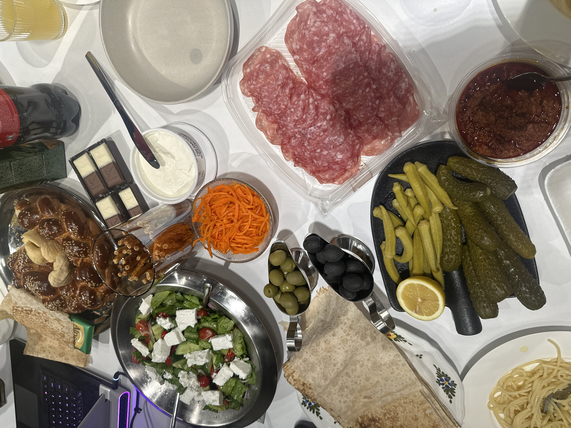 The image captures a well-lit, top-down view of a food-laden table. The scene appears to be indoors, with a white surface serving as the backdrop.
On the table, there is an assortment of food items, including:
- Sliced salami in a plastic container
- A bowl of shredded carrots
- A container of white cream cheese
- A plate of pickles, okra, and a lemon wedge
- A bowl of dark red dip
- Two small serving spoons containing green and black olives
- Several pieces of flatbread
- A plate of salad with feta cheese, tomatoes, and lettuce
- A braided sweet bread roll, topped with nuts and sesame seeds
- A box of chocolate pieces
- A plate of spaghetti
There are other objects visible in the shot, including a bottle of Coca-Cola, a couple of empty plates, a laptop, and a glass.
Without further information, it is not possible to confirm the location as Yerevan, Armenia.