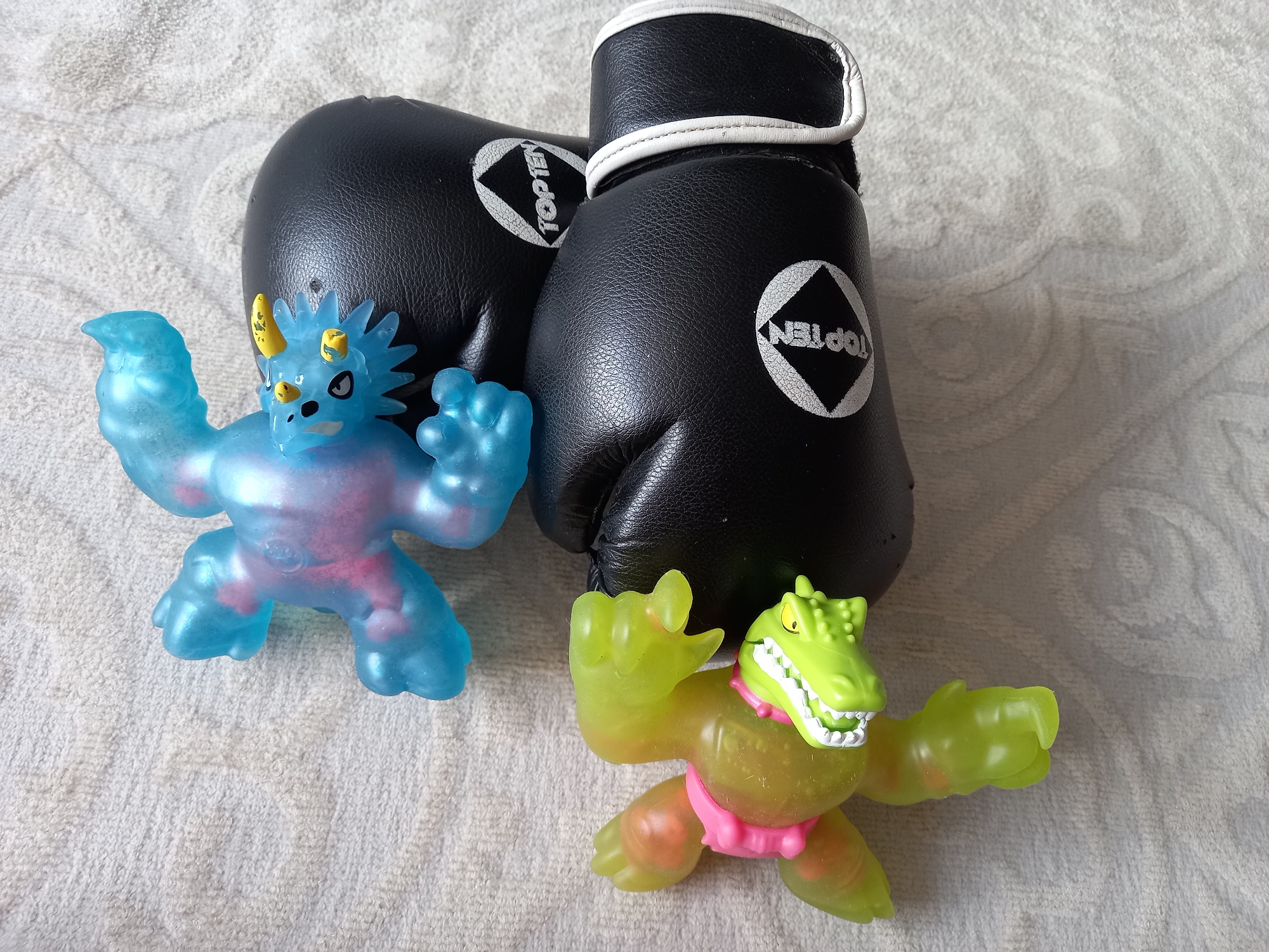 On a white textured surface, two black boxing gloves with white trim are positioned behind two stretchy toy figures. The left figure is translucent blue with a pink interior, resembling a dinosaur. The right figure is translucent green with a pink interior, resembling a crocodile. The gloves bear the brand "TOP TEN". The location is Unknown, Unknown.