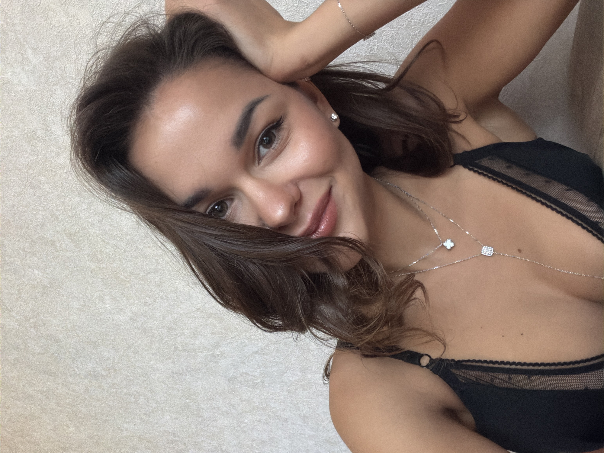 A medium shot of a woman with brown hair and light brown eyes smiling at the camera. She is wearing a black bra with lace detailing and two silver necklaces, one with a clover pendant and another with a square pendant. She is also wearing a silver bracelet. Her left hand is raised to her head, with her fingers touching her hair. The background is a textured off-white wall. There is no discernible information to suggest the location is Krasnoyarsk, Russia.