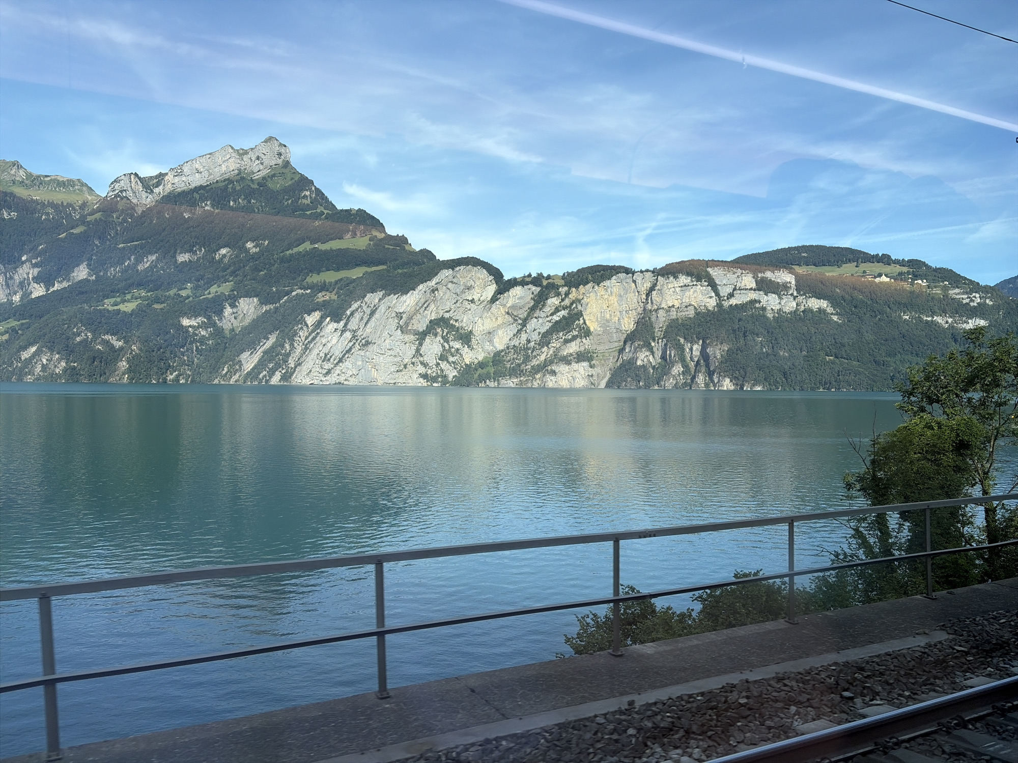 This image captures a scenic view from Morschach, Switzerland. A serene, turquoise lake dominates the foreground, its surface rippled by a gentle breeze. Along the lake's edge, a grey metal railing runs horizontally across the frame, suggesting a walkway or viewing platform. Beyond the railing, lush green trees line the shore, their reflections faintly visible on the water. In the background, imposing mountains rise steeply, their rocky faces contrasted with verdant slopes covered in trees and patches of grass. The sky above is a vibrant blue, streaked with wispy white clouds and a faint contrail from an aircraft. The overall scene conveys a sense of natural tranquility and the majestic beauty of the Swiss landscape.