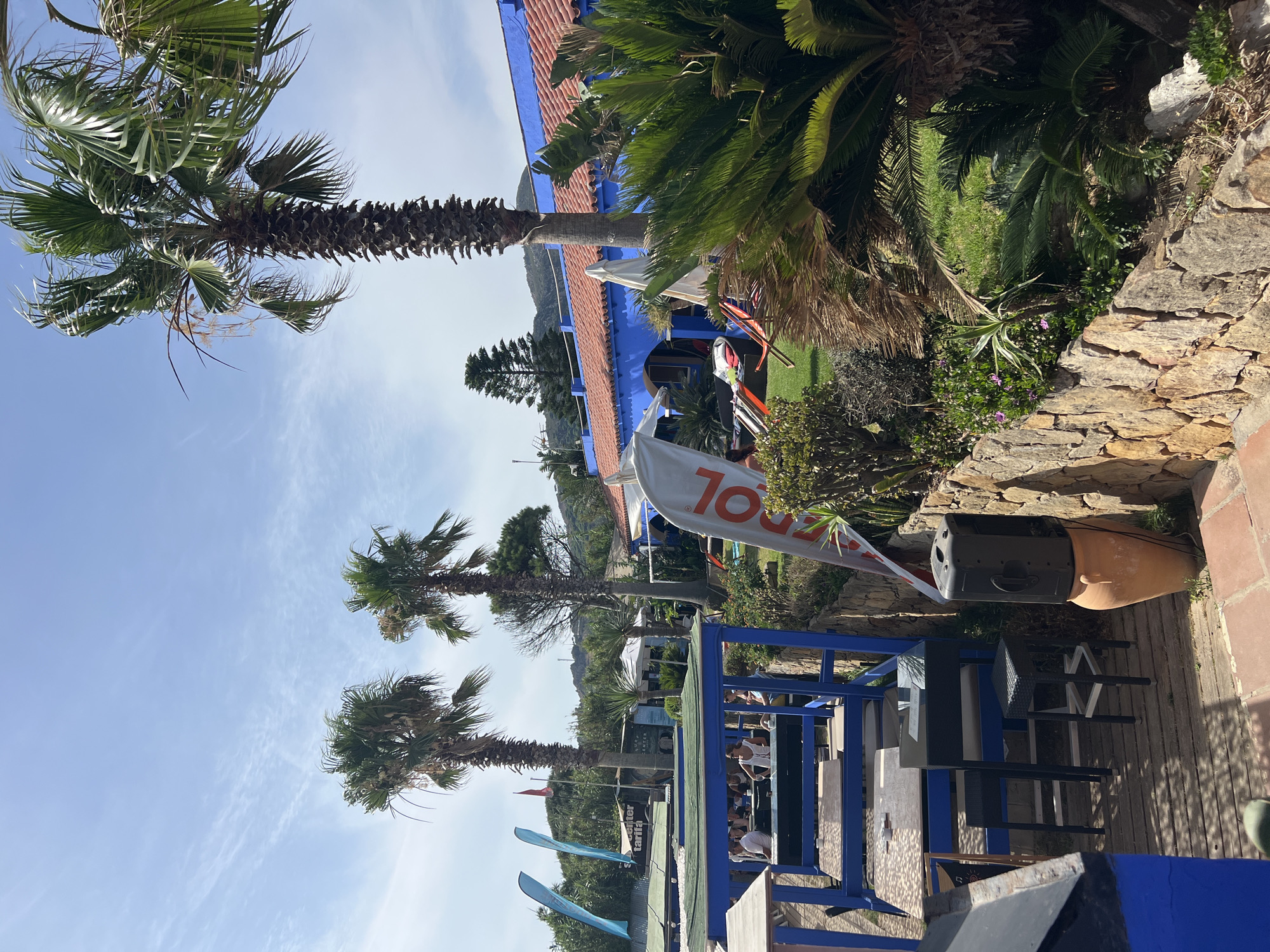 The image depicts an outdoor seating area at a beachside establishment in Tarifa, Spain, on a sunny day. Palm trees are prominent throughout the scene, framing the view of a blue building with a red tiled roof. In the foreground, a wooden deck with blue railings holds tables and chairs, where several people are seated, some appearing to be dining. To the right, a stone wall separates the seating area from a grassy lawn with sun loungers and umbrellas. A large banner with an orange logo and white lettering, possibly advertising a brand or event, stands near the wall. A black speaker is placed on top of a large terracotta pot. In the background, flags and structures suggestive of a watersports center are visible. The sky is a clear blue with scattered wispy clouds.