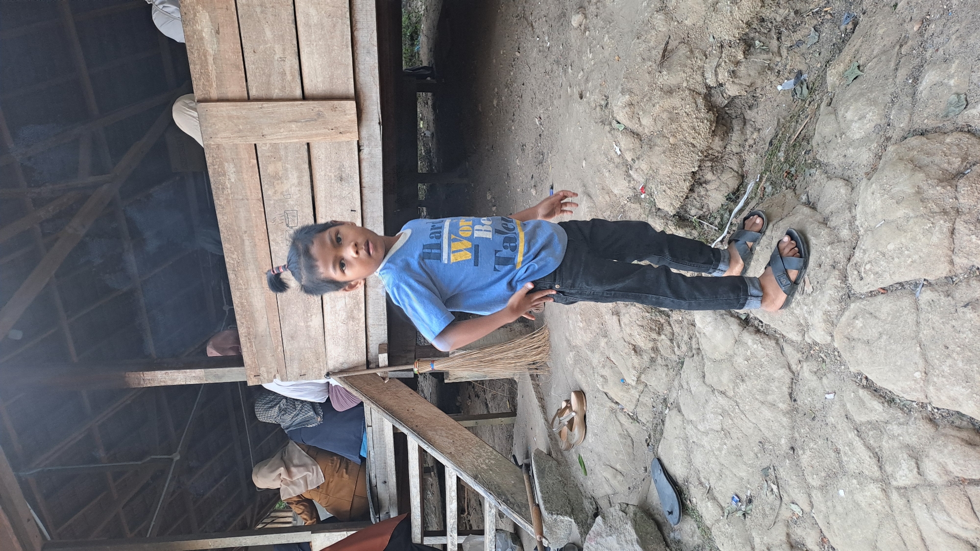 A young boy with his hair tied in a bun stands in front of a wooden structure in Geulanggang Baro, Indonesia. He is wearing a blue t-shirt with yellow and white lettering and black pants. The boy is standing on a rocky surface. To his left, a broom leans against a wooden ramp leading up to the structure, where several people are seated. The ground is uneven and appears to be a mix of dirt and rocks.