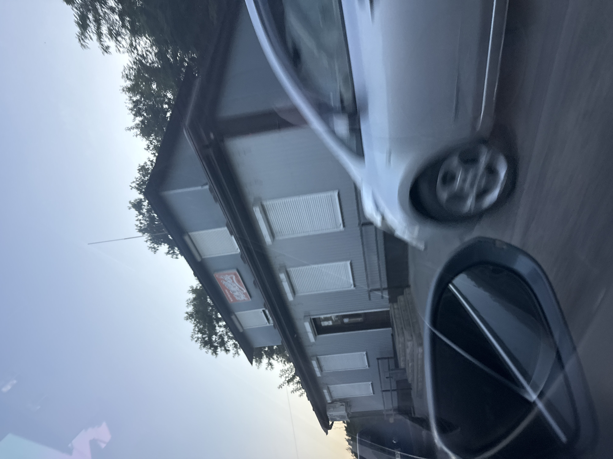 In the city of Orlivka, Ukraine, a one-story building with a gray exterior and a dark roof stands under a clear sky. The building has several windows with white roll-up shutters. A red sign with white text is mounted on the upper part of the building's facade. To the right of the building, a silver car is in motion, its wheels blurred by speed. In the foreground, the dark side mirror of a vehicle reflects the sky and trees. The scene appears to be captured from another moving vehicle, suggesting a street view.