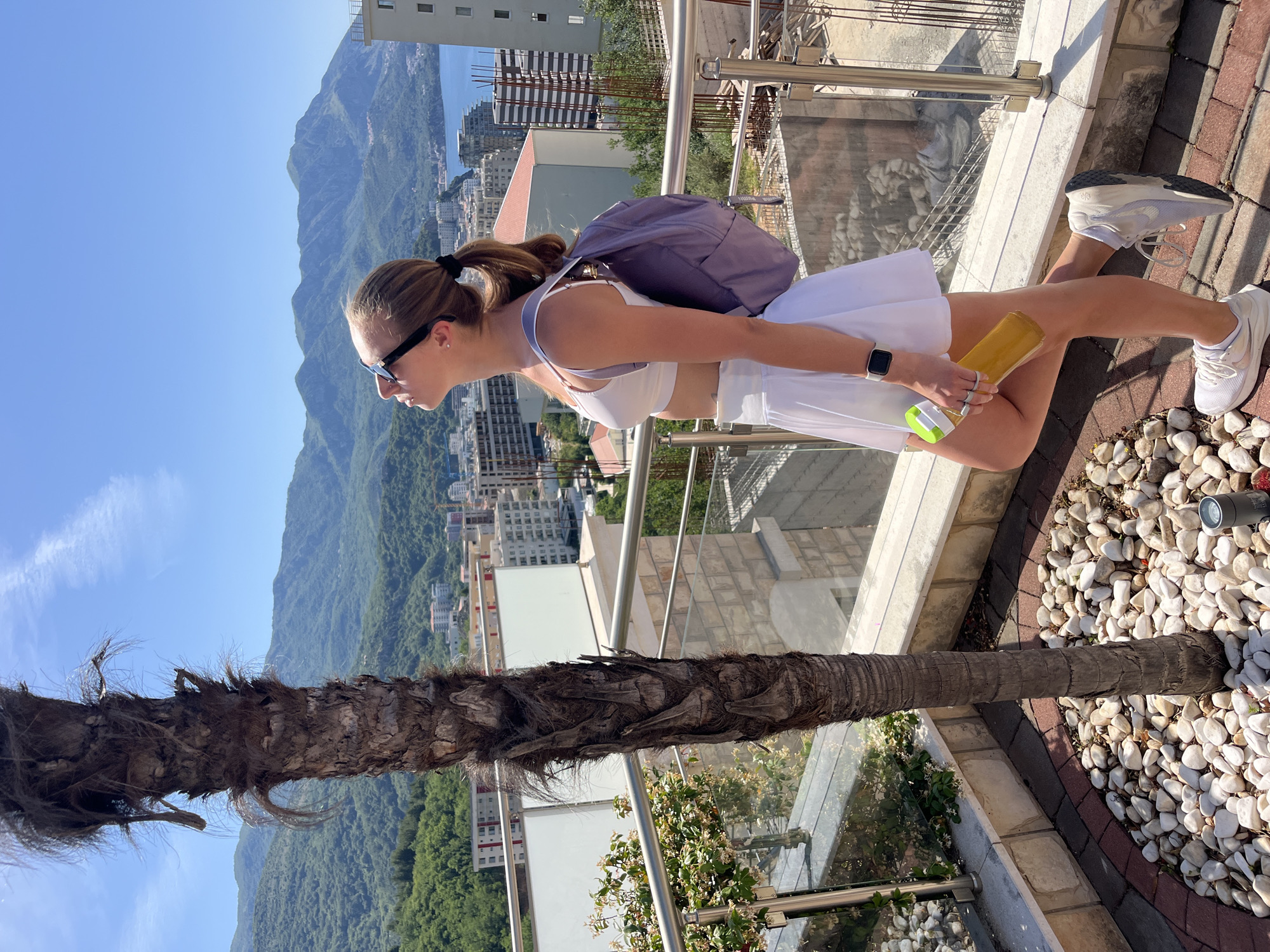 A young woman stands on a patio in Boreti, Montenegro, with a backdrop of mountains, buildings, and the sea. She wears sunglasses, a white sports bra and skirt, and white sneakers. A purple backpack is slung over her shoulder. In her hands, she holds a yellow beverage container. The patio is decorated with white rocks and bordered by a stainless steel railing. A palm tree trunk is partially visible in the foreground.
