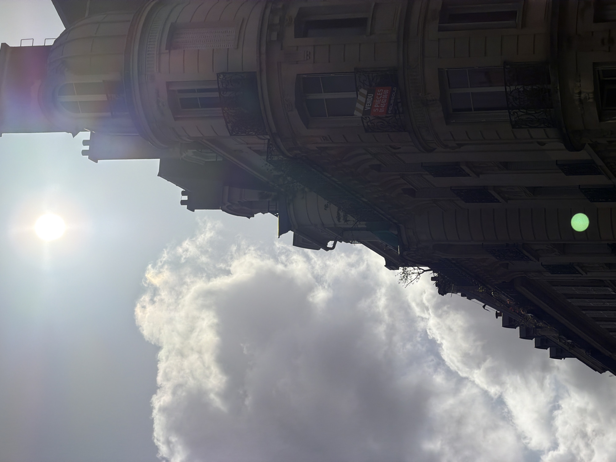 The image shows a Parisian building on a partly cloudy day. The building has multiple windows with wrought iron balconies. A "VENDU 5 PIECES" (SOLD 5 ROOMS) sign is visible on one of the windows, likely indicating that the apartment has been sold. The sky is visible in the lower half of the image, with some clouds partially obscuring the sun. The image appears to have been taken from a low angle, looking upward at the building. The location is Paris, France, based on the architectural style of the building and the type of sign displayed.
