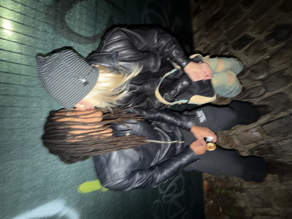 Two individuals are positioned on a concrete surface, possibly a floor or wall. One person has long dreadlocks, a black leather jacket, and dark pants. Their companion has blonde hair, a striped beanie, and a black leather jacket. They are holding a purse. The backdrop suggests a dark, possibly urban environment. One person appears to be holding something gold in their hands.