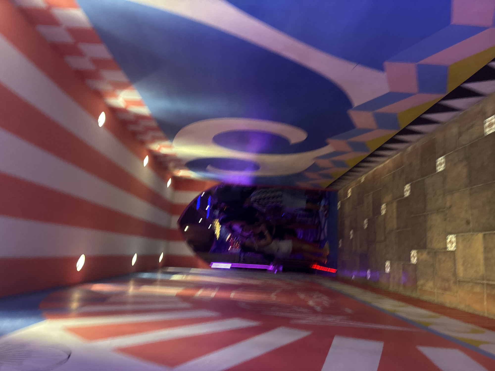 This media file depicts an indoor scene with a walkway that has a striped red and white pattern on the floor and walls. The ceiling is decorated with a blue pattern that includes stylized curved shapes and what appear to be three-dimensional cubes. Along the walls, there are small, oval-shaped lights. The walkway leads into a darker area with a curved structure that houses people and is illuminated with colorful lights, including purple and red.
The overall impression is of an entertainment venue or a themed area, possibly an amusement park or a carnival, given the vibrant colors and decorative patterns. The motion blur suggests that the image was taken while moving, possibly from a ride or while walking through the space. The activity taking place seems to be people congregating within the illuminated curved structure. There is no visible text in the image. The lighting and atmosphere suggest it could be evening or an indoor location with artificial lighting. The location context provided is North Kuta, Indonesia.