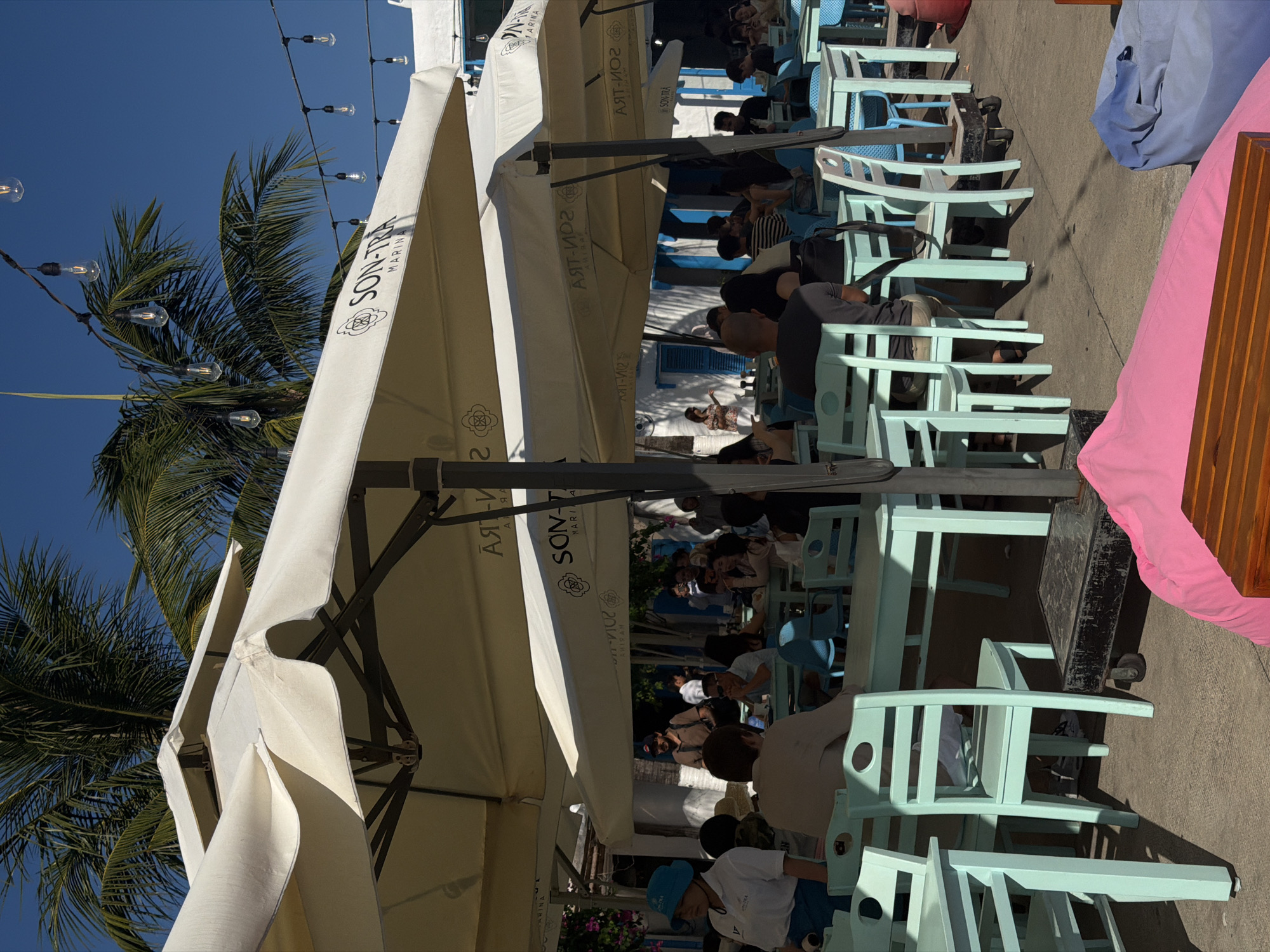A photograph, tilted at a ninety-degree angle, features outdoor seating beneath large umbrellas, likely at a restaurant or cafe. The setting seems to be in the city of Đà Nẵng, Vietnam.
Visible are tables and chairs, mostly painted a light turquoise color, suggesting an open-air dining area. People are seen seated at some of the tables, interacting with each other. Above, large white umbrellas provide shade. The words "SON-TRA MARINA" are inscribed on the umbrellas.
In the background, one can see a partly cloudy sky and palm trees, indicating a coastal location. String lights are strung between the trees and umbrellas, adding to the atmosphere.
The scene depicts a normal daytime activity of people enjoying the amenities of a restaurant in a coastal environment. The tilt of the image affects the composition, but the key elements are clearly identifiable.