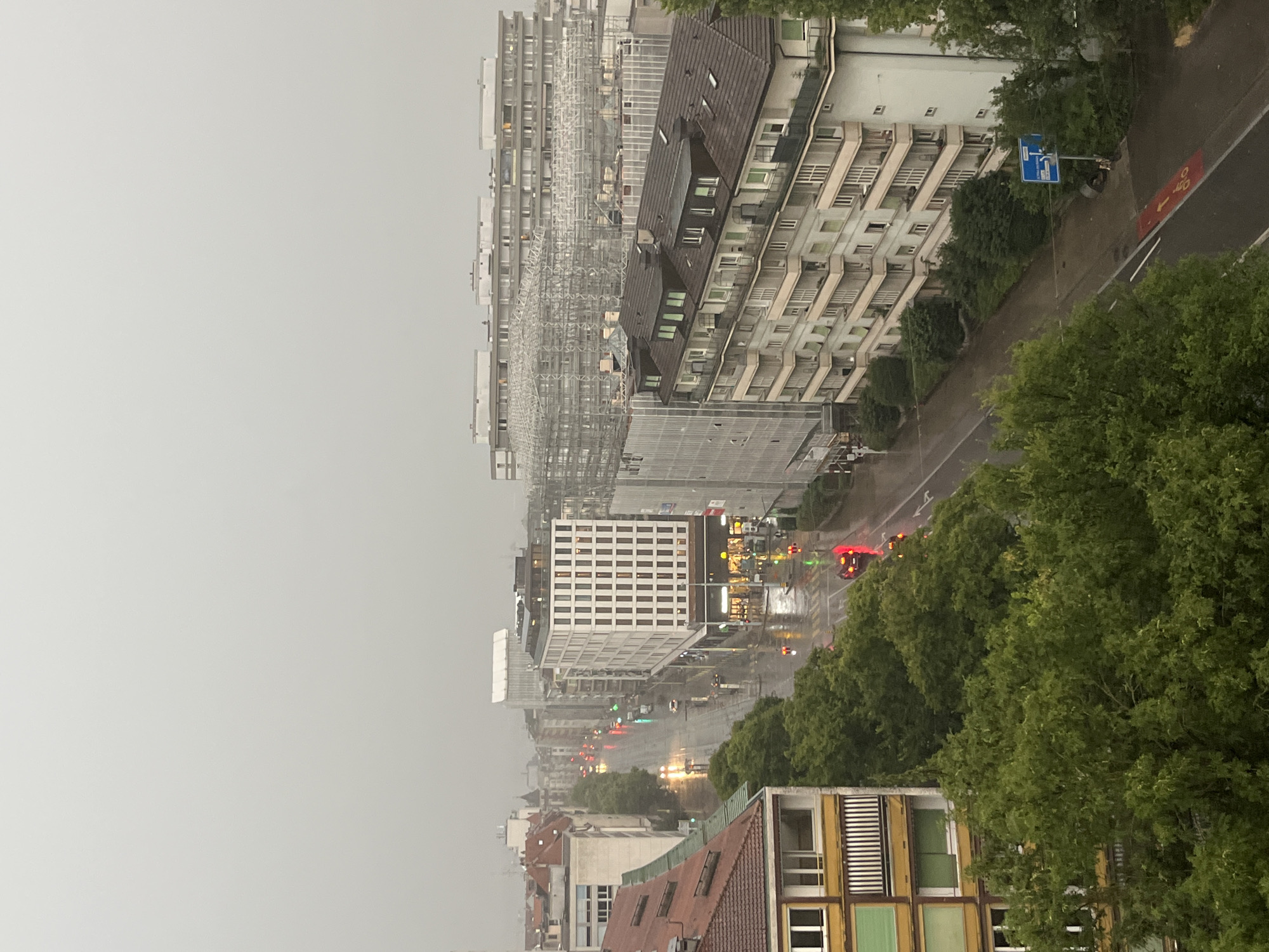 This is a high-angle, medium shot of a street in Geneva, Switzerland, during a rainy day. The sky is overcast and gray, and the street and sidewalks are wet, reflecting the lights from the buildings and traffic signals.
On the left side of the street, there are several multi-story buildings. The closest building has a reddish-brown tiled roof and a facade with yellow and green accents. Further down the street, a modern white building with many windows stands out. Across the street, a pale pink building with multiple balconies is visible.
Vehicles, including cars and possibly a tram, are visible on the wet street, their headlights and taillights creating streaks of light. Traffic signals are illuminated with red and green lights. There are also several trees lining the street, their leaves appearing dark and wet from the rain.
In the background, more buildings are visible, some with scaffolding, suggesting ongoing construction or renovation. A blue street sign with white symbols is present on the right side of the road. A red painted area on the road indicates a bicycle lane. The overall impression is of a typical urban scene during a downpour.