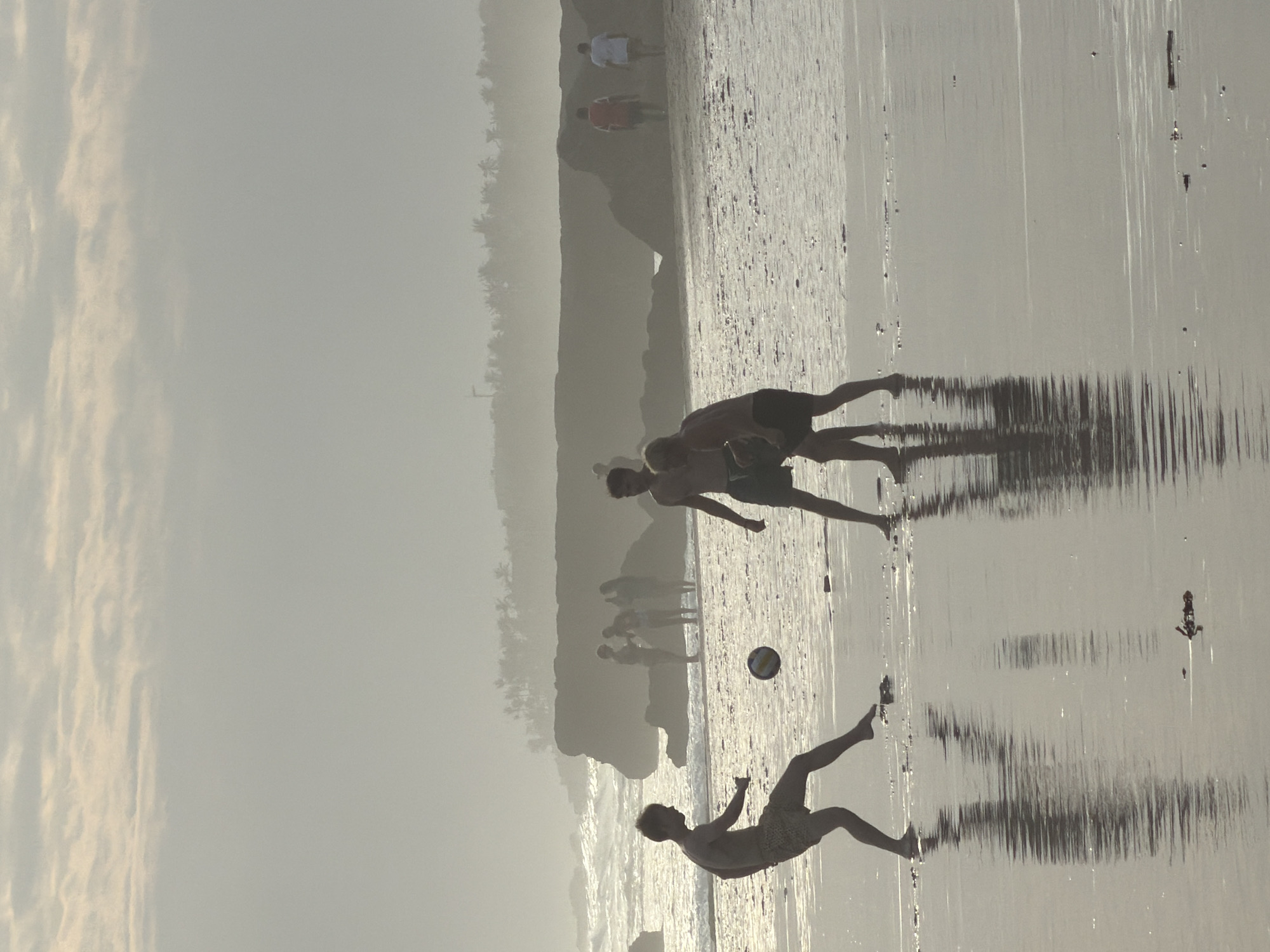 In North Kuta, Indonesia, at sunset, three figures are silhouetted on a beach as they play soccer. The scene is bathed in soft, hazy light, with the sky filled with muted clouds. The foreground features wet sand reflecting the figures and the sky. In the mid-ground, two figures are actively engaged in playing soccer with a ball, while a third figure is observing. Behind them, a line of dark rocks forms a natural barrier, and beyond that, more silhouetted figures are seen near the water's edge. The background consists of the ocean with gentle waves and a distant, hazy shoreline with palm trees.