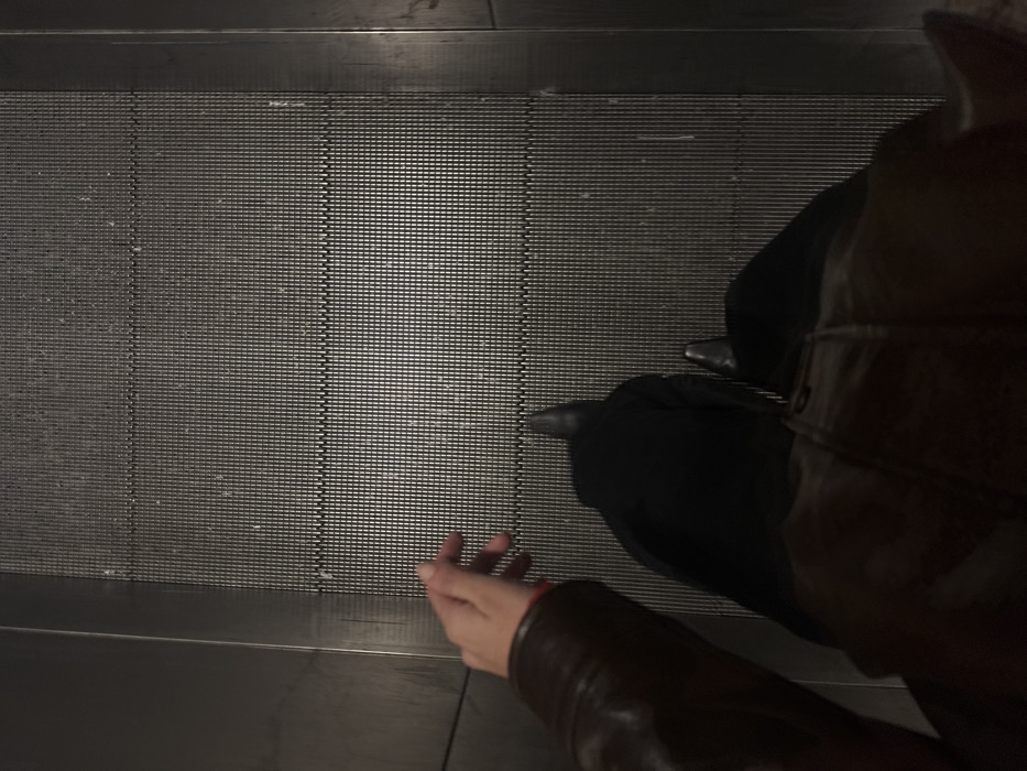 A single individual is depicted from a first-person, downward perspective, standing on a moving walkway or escalator within an indoor, artificially lit environment. The subject wears dark trousers, black pointed-toe shoes, and a brown jacket. The left hand is extended forward, fingers slightly separated. The walkway surface comprises ridged metallic panels, bordered by smooth, dark vertical side panels. This infrastructure is consistent with a public transit facility, such as a metro station or airport concourse, commonly found in Paris, France.