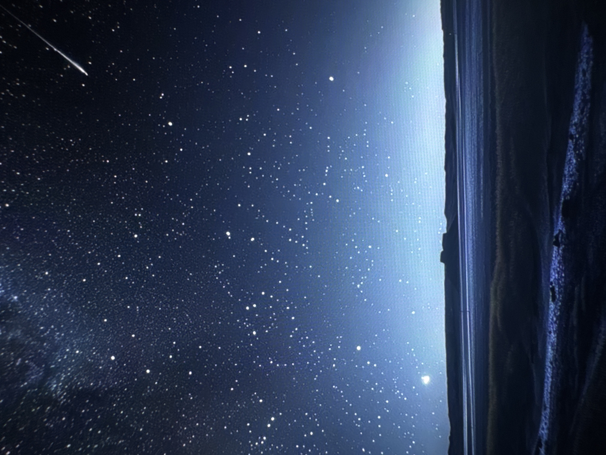 A wide-angle, eye-level shot captures a vast expanse of a star-filled night sky above a dark, silhouetted landscape. The sky is a deep blue, transitioning to a lighter, almost luminous blue towards the horizon, filled with countless white stars of varying sizes. A single, bright shooting star streaks across the upper right portion of the frame. In the distance, a flat, dark landmass with irregular, layered formations forms the horizon. Below this landmass, a dark, reflective surface, possibly a body of water or a dry lakebed, stretches across the midground, with faint horizontal lines suggesting ripples or reflections. In the foreground, a dark, textured area of dry grass or brush is visible, with a patch of lighter, possibly wet or frost-covered ground in the lower left corner. The overall scene is serene and remote, with no visible signs of human activity. The image appears to be taken at an outdoor location, likely in a rural or wilderness setting, under a clear night sky.