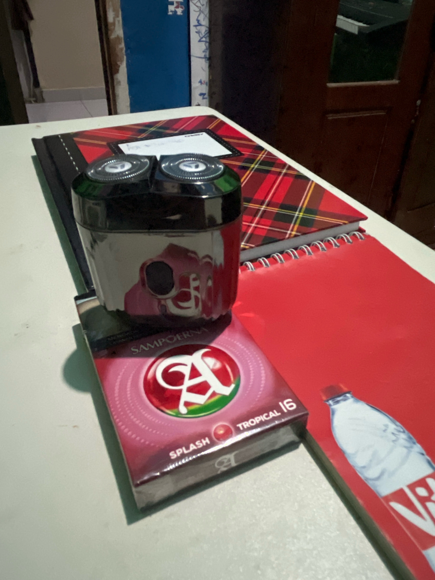 The image shows a close-up, eye-level shot of a table with several items arranged on it. The primary subjects are a black electric razor with two circular heads, a pack of "Sampoerna Splash Tropical 16" cigarettes, a red spiral-bound notebook, and a partially visible bottle of "Vit" sparkling water. The razor is placed on top of the cigarette pack. Behind the razor and cigarette pack, there is a black notebook with a red and black plaid cover. The red notebook is positioned in front of and to the right of the plaid notebook. The Vit bottle is on the right side of the frame, lying on its side on top of the red notebook, with its cap facing left. The background is blurred, showing a wooden door on the right and what appears to be a doorway or opening with a blue painted wall on the left. The lighting suggests an indoor setting, possibly a room. There are no people or animals visible in the image. The scene is composed in a way that emphasizes the arrangement of the objects on the table. The exact location in Talang Kelapa, Indonesia, cannot be determined from the visual information alone.