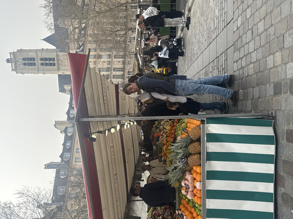 An outdoor fresh produce market is depicted on a cobbled street in the Saint-Germain-des-Prés neighborhood of Paris, France, on an overcast day. In the foreground, a market stall, skirted with green and white vertical stripes, displays an assortment of fresh fruits including pineapples, oranges, limes, and other items. The stall is covered by a white canopy with red trim, illuminated by a string of visible light bulbs. A male vendor in a dark jacket is partially visible behind the stall on the left. To the right of the stall, a man in a blue vest and jeans holds an infant dressed in a light-colored, full-body outfit. A young child, wearing a dark jacket, stands beside the man's left leg, looking towards the fruit display. In the midground, numerous individuals are present on the cobblestone street, browsing other market stalls that extend into the distance. A woman pushes a stroller further down the path. The background features classic Parisian buildings with multiple windows and mansard roofs, interspersed with bare trees. Dominating the far background on the right is the distinctive square bell tower of the Church of Saint-Germain-des-Prés, topped with a clock.