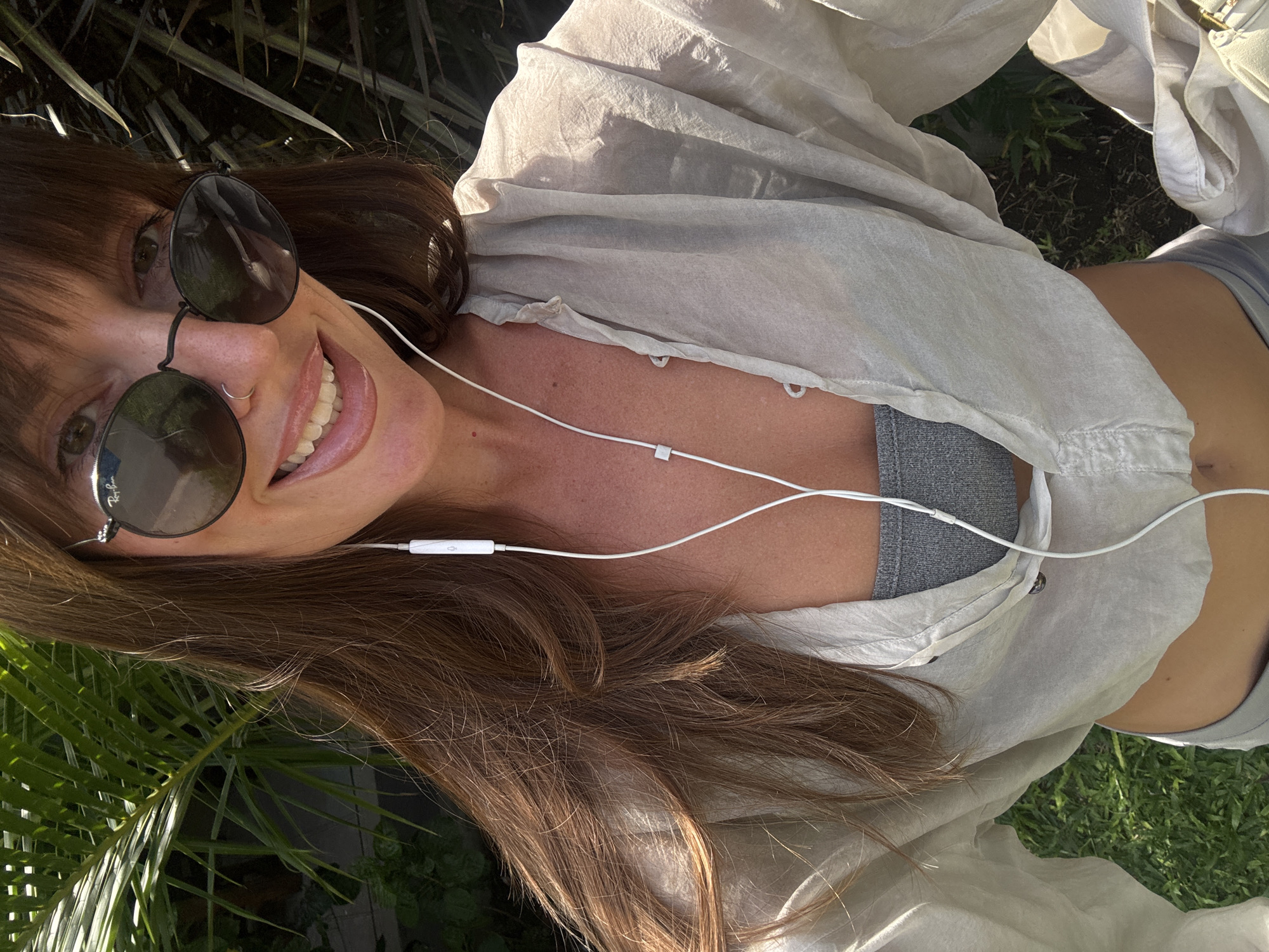 A woman lies on a patch of grass, likely in North Kuta, Indonesia, judging from the lush, tropical foliage in the background. She wears round, dark sunglasses and a light-colored, button-down shirt open at the chest, revealing a grey sports bra. White wired earphones are plugged into her ears. She is smiling. Her long, brown hair is tousled around her.