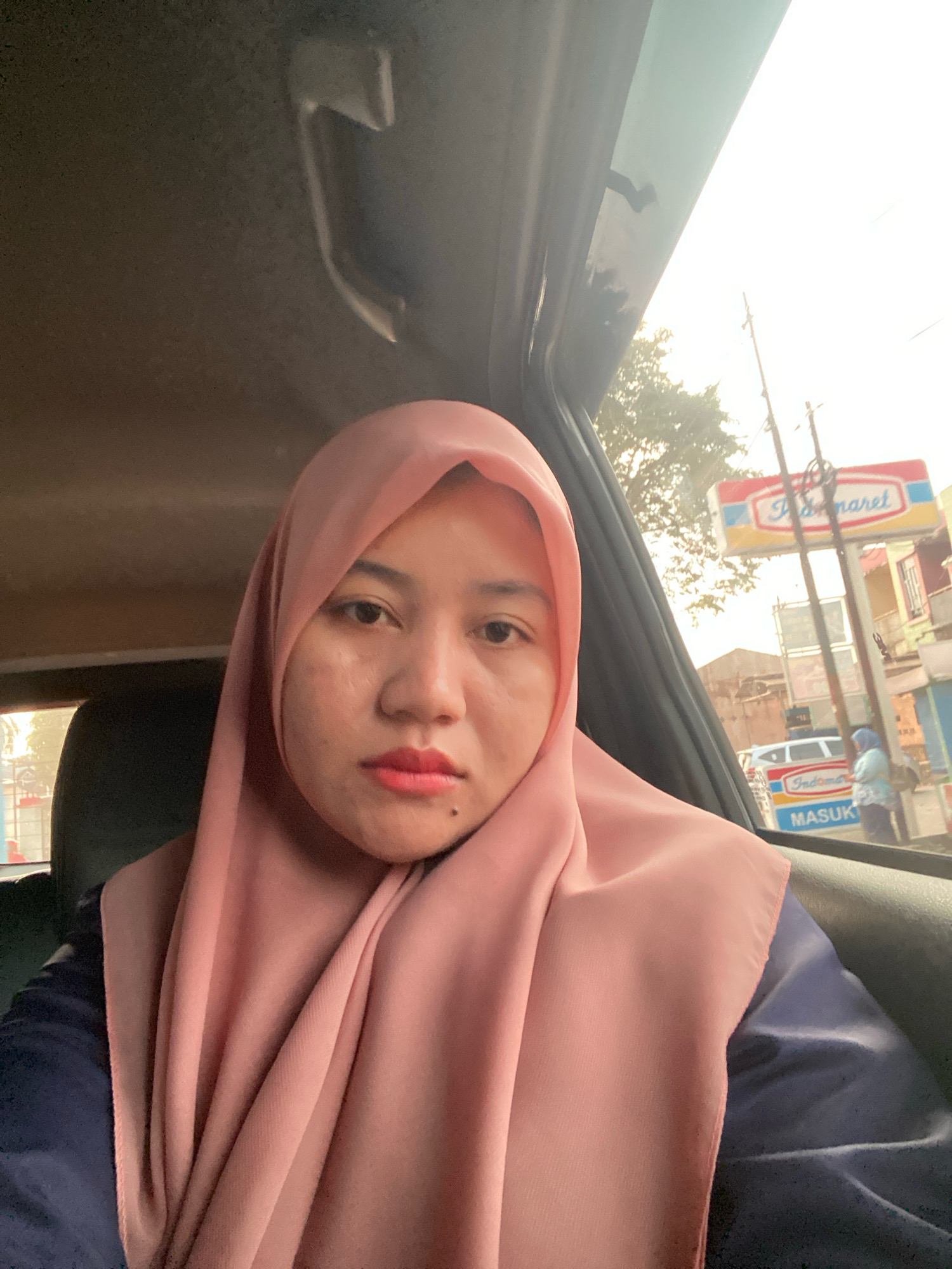 A woman wearing a pink hijab and a navy blue shirt is inside a car, taking a selfie. The woman has fair skin, dark eyes, and is wearing pink lipstick. A mole is visible on her chin. The car's interior is dimly lit, with a visible handle on the ceiling. Through the car window, a street scene in Banjarbaru, Indonesia, is visible. A sign for "Indomaret" is prominent, along with utility poles and other buildings. The sky is bright and overcast, suggesting it is daytime. A person wearing a light blue hijab is standing on the street. The overall lighting suggests it is either early morning or late afternoon.