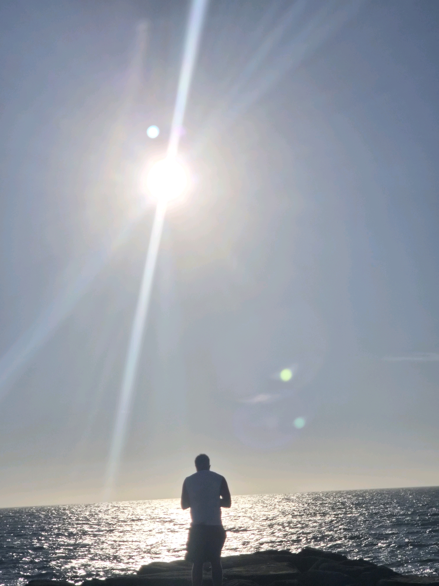 A lone male figure stands on a rocky outcrop, facing away from the camera, towards a sun-drenched ocean. The sun, high in the sky, casts a bright, elongated lens flare diagonally across the frame, and two smaller, circular green lens flares are visible in the upper right quadrant. The sea is choppy, with sunlight glinting off the waves, creating a shimmering effect. The man is wearing a light-colored t-shirt and dark shorts. The scene suggests a moment of solitary contemplation by the sea. The location is the coastline of Esposende, Portugal, known for its dramatic rocky shores and expansive ocean views.