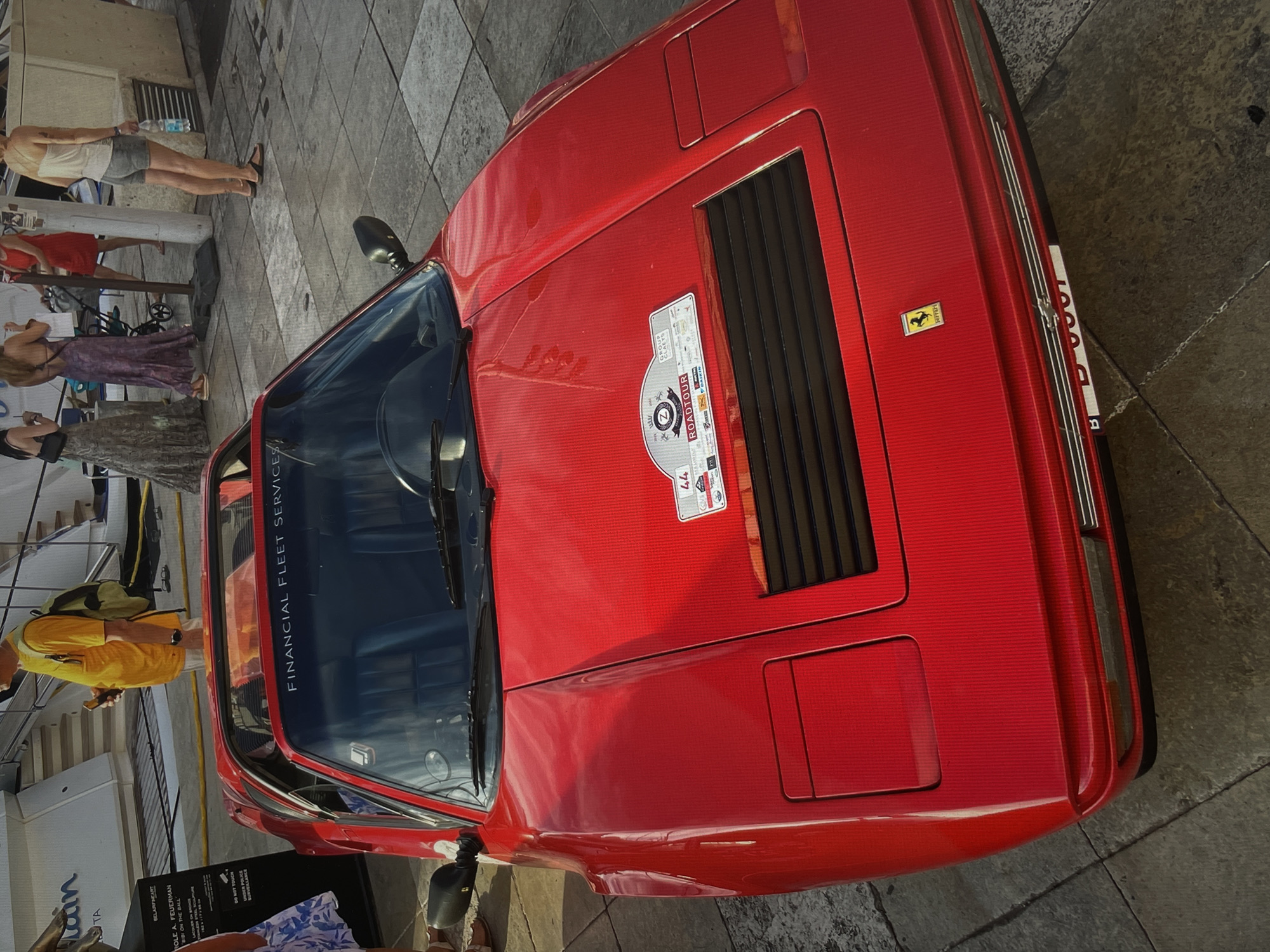 The image shows a red Ferrari car parked in an urban area, likely in the city of Unknown, Unknown. The car has a sticker on the hood related to a "Roadtour" event and the words "FINANCIAL FLEET SERVICES" are visible on the windshield. A group of people are standing around the car and in the background, including one person wearing a yellow shirt and a backpack. A sign related to "BELARFINEART" is visible near the bottom of the frame. The ground is paved with square stone tiles.