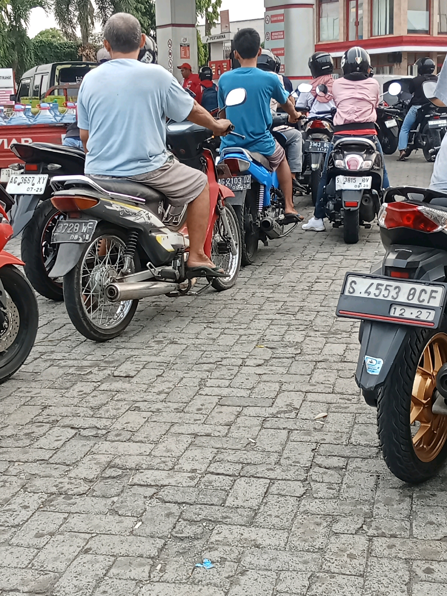 In the city of Jelakombo, Indonesia, this image captures a scene at a gas station with a group of people on motorcycles. The foreground is dominated by the rear of a motorcycle with a license plate reading "S.4553 BCF" and "12.27." To its left, another motorcycle's wheel and part of its license plate are visible. In the middle ground, several motorcycles and their riders are lined up, with some waiting. One rider, wearing a blue t-shirt, is facing away from the camera. Behind him, other riders are also seated on their motorcycles. The background reveals more motorcycles and a gas station with pumps and buildings. The ground is paved with interlocking grey bricks.