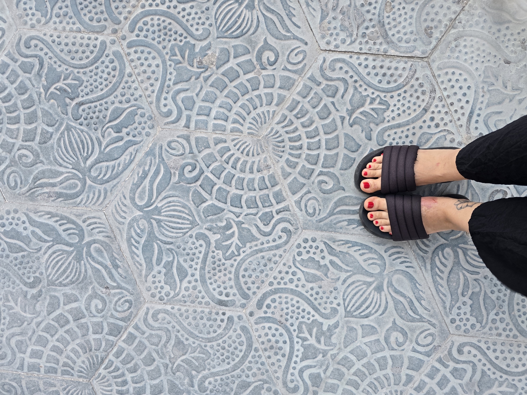 A top-down, medium shot shows a pair of feet wearing black slides with red toenails, standing on hexagonal tiles with a blue-grey, embossed floral and geometric pattern. The tiles are laid in a honeycomb arrangement, and the pattern varies between hexagonal sections. A portion of black fabric, likely clothing, is visible in the lower part of the frame. The scene appears to be an outdoor pedestrian area.