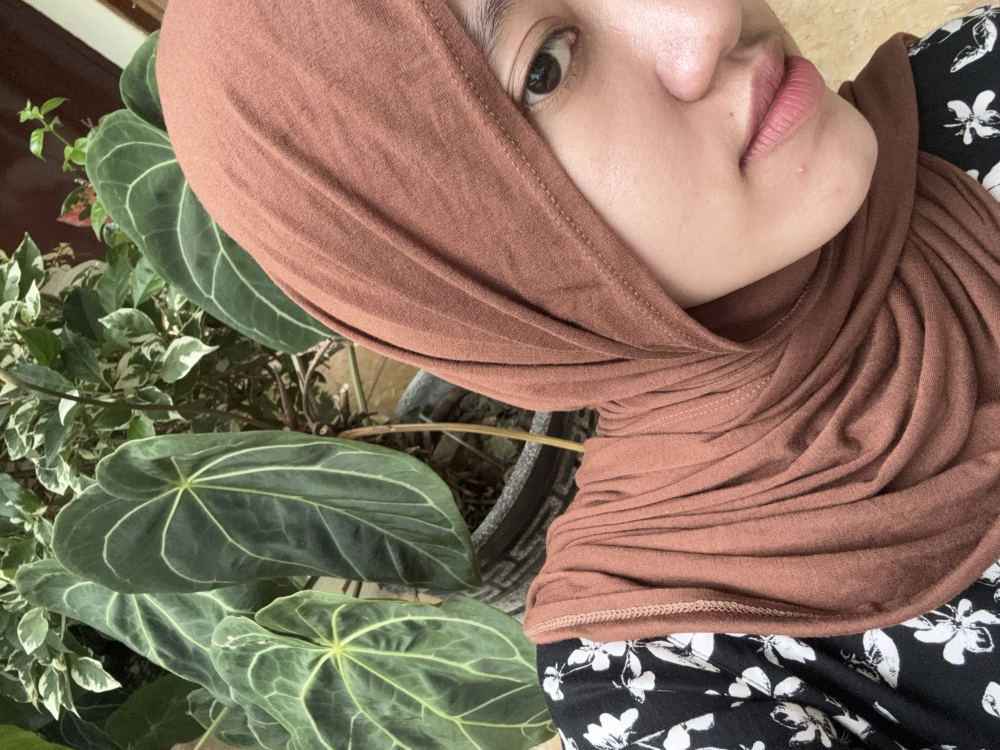 A person wearing a brown hijab is positioned near foliage. The person is wearing a black and white floral patterned shirt. The foliage contains large green leaves with white veining. The location is outdoors with a partially visible dark brown wall in the background. The scene is potentially located in Gadingrejo, Indonesia, based on the prompt.
