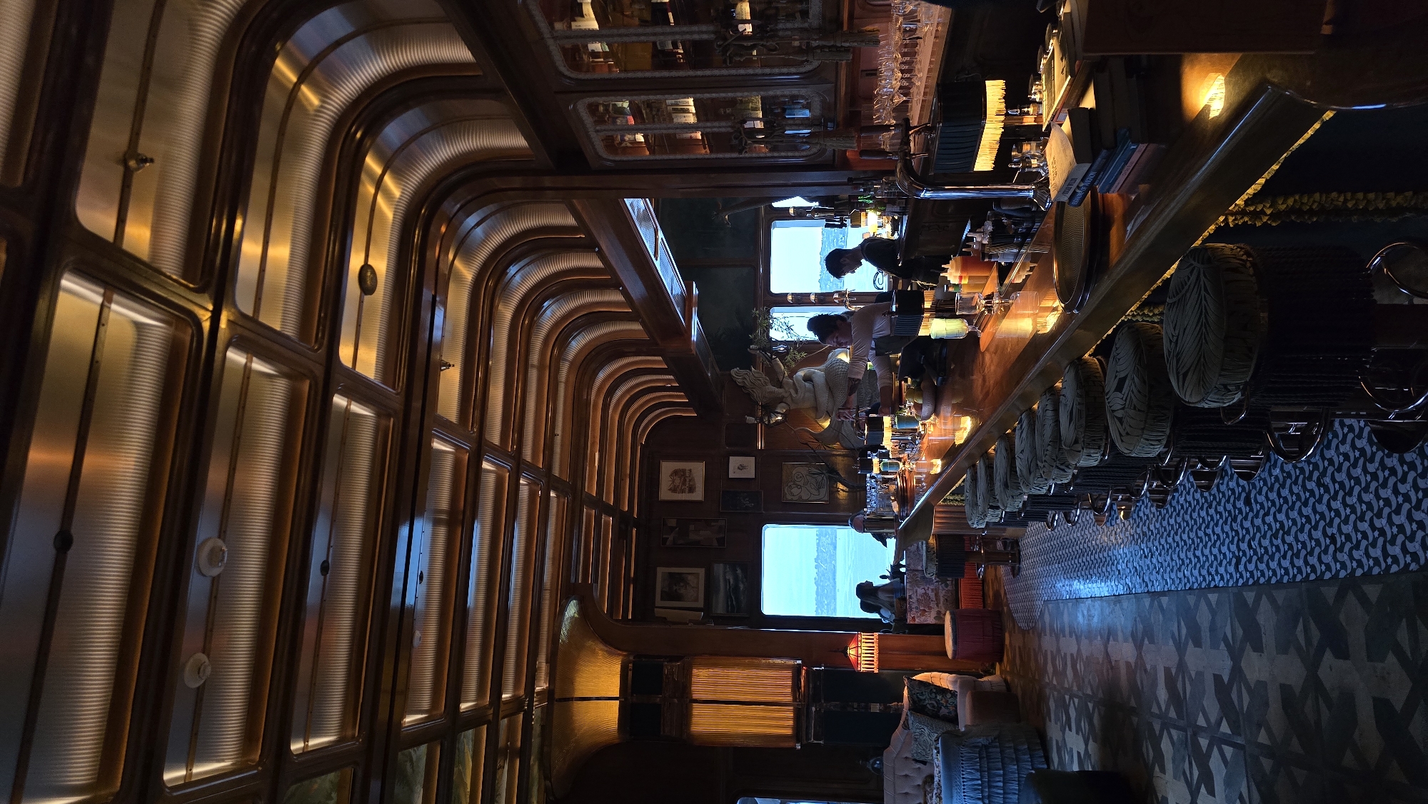 The image depicts the interior of a dimly lit bar with a prominent wooden bar counter stretching across the frame. The bar is lined with plush, patterned bar stools. Behind the counter, bartenders are visible preparing drinks, and bottles of liquor are neatly displayed on shelves. A large window behind the bar offers a view of a body of water and a distant shoreline, suggesting a waterfront location. The ceiling features a distinctive design with ribbed, translucent panels illuminated from within, casting a warm glow. The overall ambiance is sophisticated and cozy, with art pieces adorning the wood-paneled walls. The location is identified as a bar in San Diego, United States of America, likely a high-end establishment with a focus on its views and atmosphere.