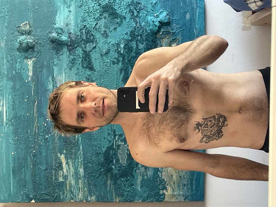 An adult male, estimated to be between 30 and 40 years old, is shown from the chest up, taking a mirror selfie. He has light brown hair, blue eyes, and visible chest hair. A black and grey tattoo depicting a stylized animal head (possibly a wild boar or similar creature) entangled with thorny branches is present on his left rib cage. He holds a black smartphone, featuring multiple rear camera lenses, in his right hand, which partially obstructs his upper chest. Behind him, a large, rectangular abstract painting dominates the wall. The painting is primarily composed of textured, cracked-effect strokes in various shades of blue and turquoise, with some raised elements and scattered white speckles, suggesting an oceanic or weathered theme. The wall itself is a plain, light color. A narrow, light-colored surface is visible at the bottom of the frame, and a corner of a blue and white checkered fabric is present on the bottom right. The specific location within Budva, Montenegro, is not discernible from the visual information provided.